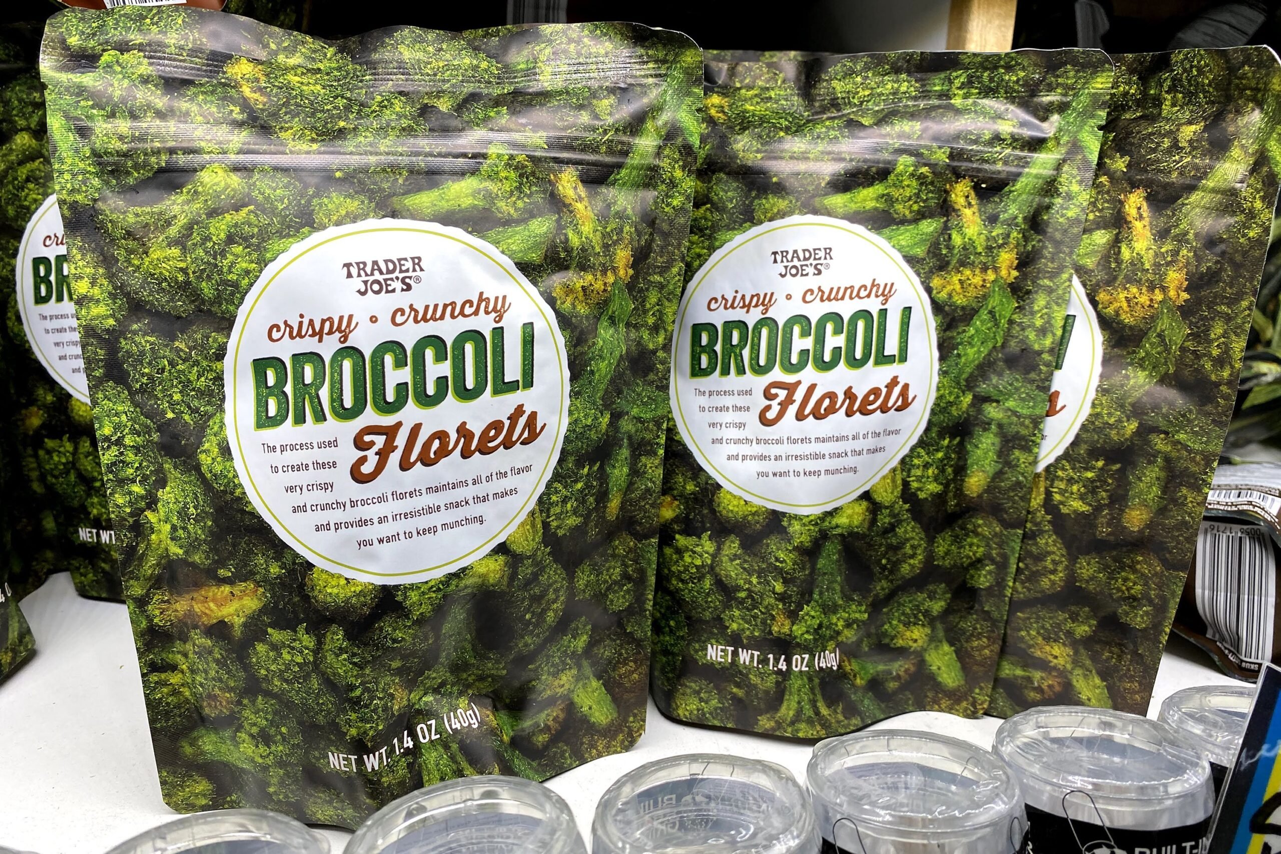 Three bags of Trader Joe's Crispy Crunchy Broccoli Florets at a Trader Joe's, on a shelf, several other bags in the background