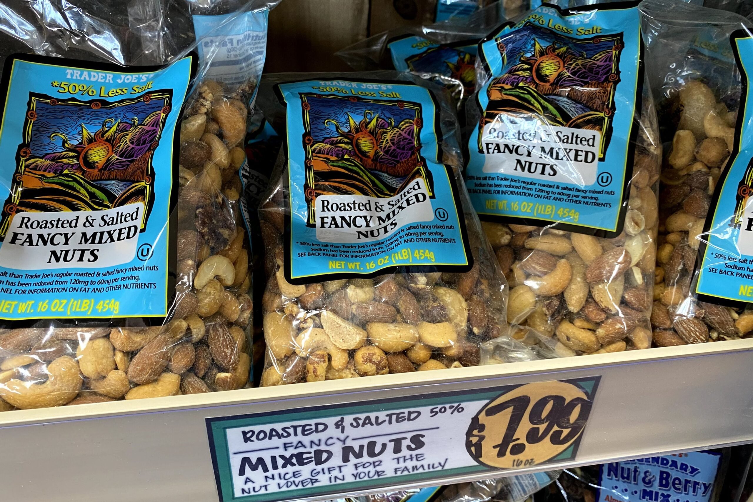 Several packages of Trader Joe's Roasted & Salted Fancy Mixed Nuts at a Trader Joe's, on a shelf, fun sign underneath