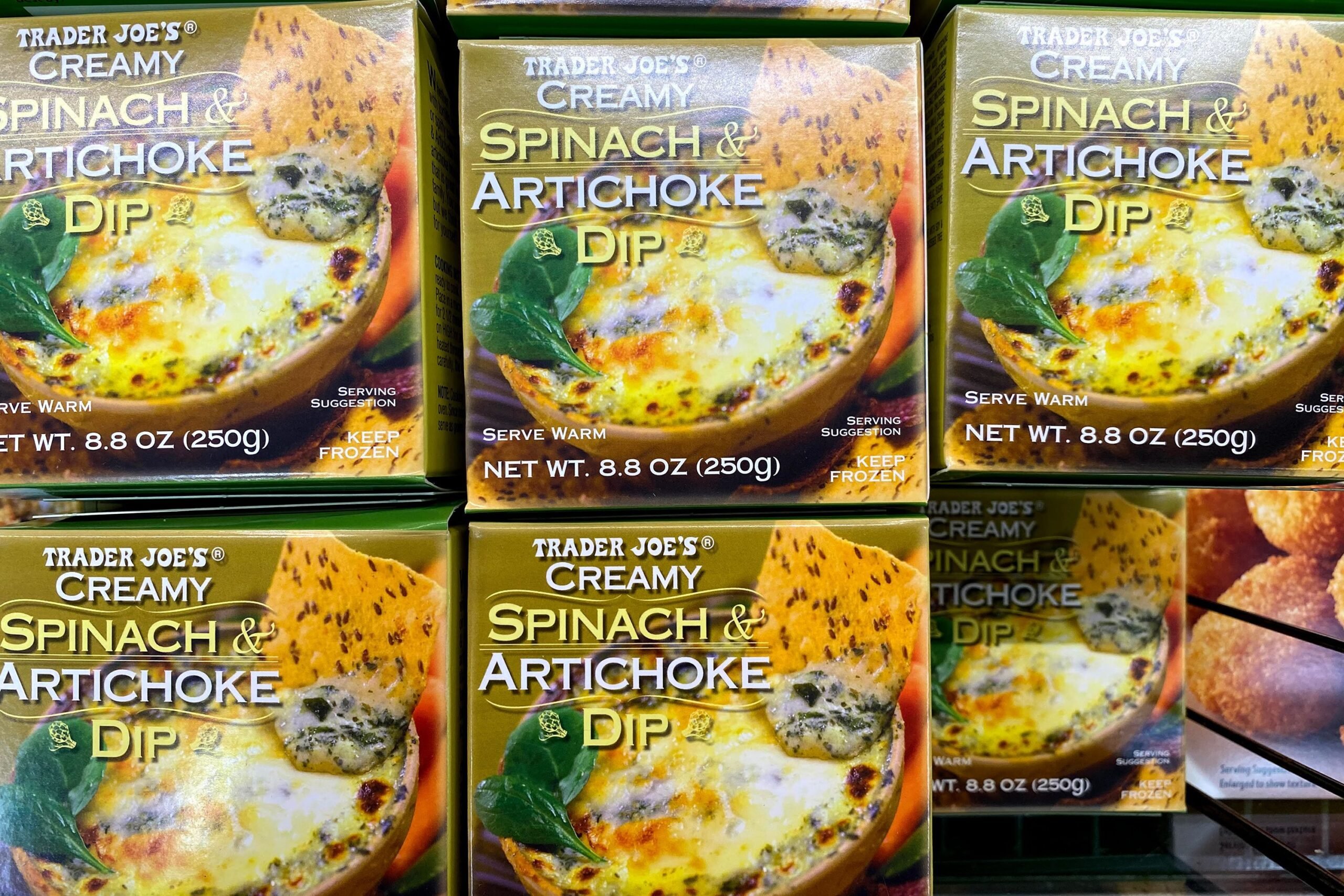 Three rows of boxes Trader Joe's Creamy Spinach & Artichoke Dip at a Trader Joe's, in a refrigerator on a shelf, one box on top of another