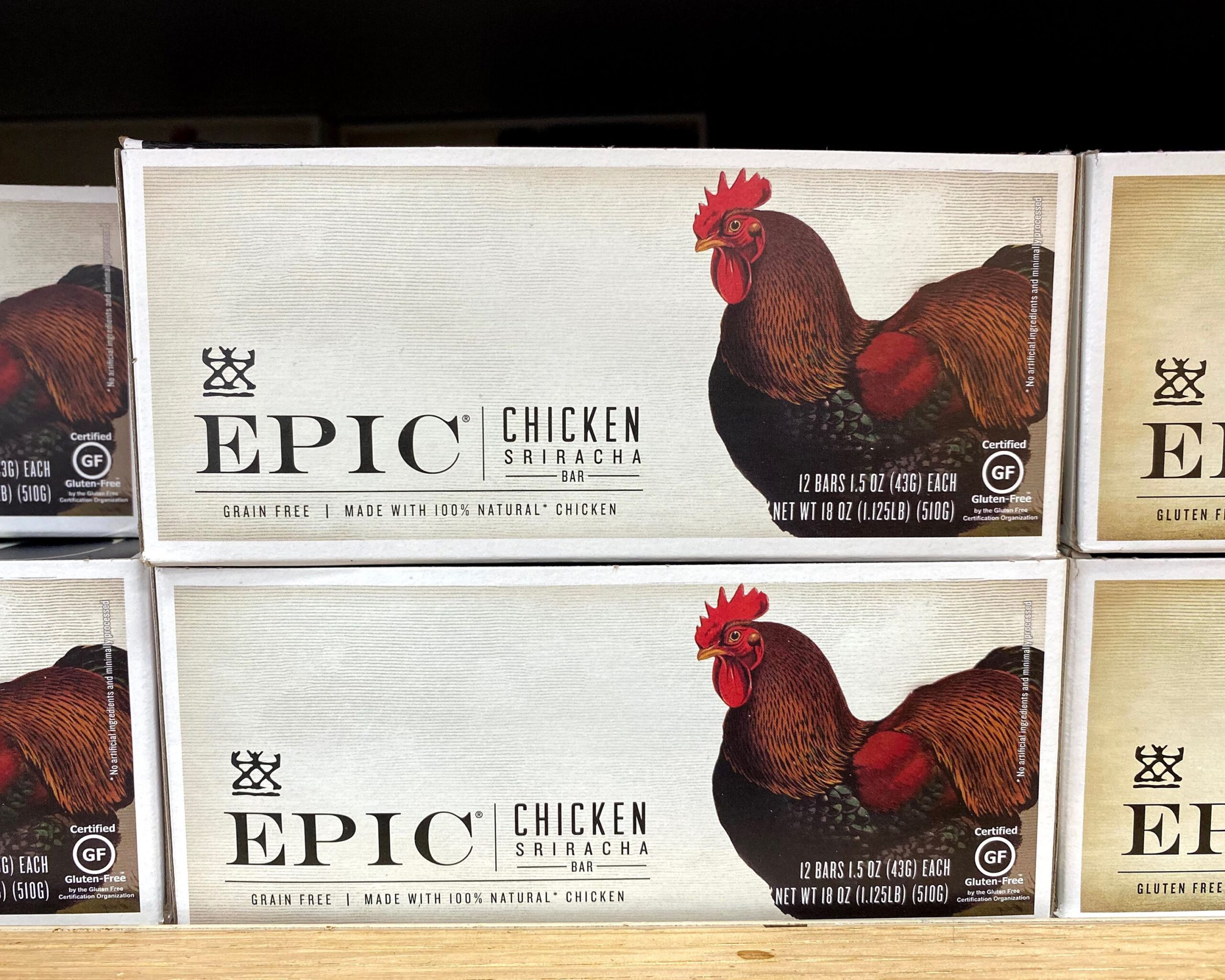 Row of boxes of Trader Joe's Chicken Sriracha Epic Bars at a Trader Joe's, on a wooden shelf, more boxes of bars on both sides