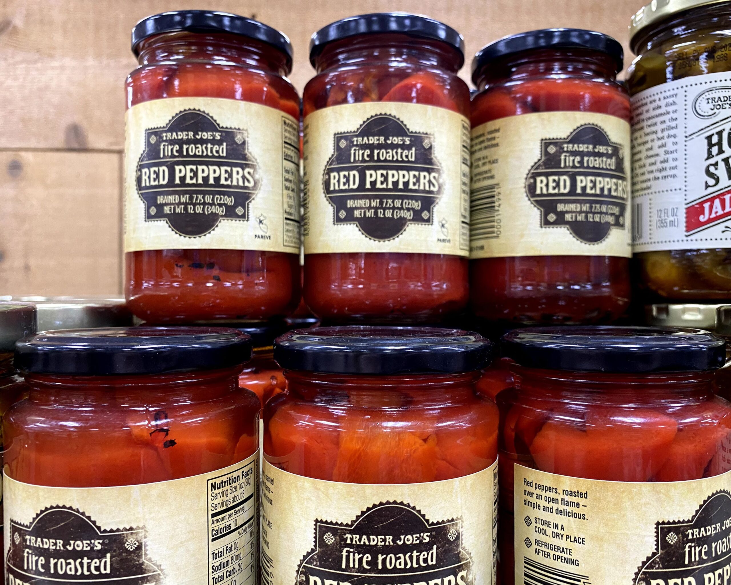 Two stacks of many jars of Trader Joe's Fire Roasted Red Peppers at a Trader Joe's, a wooden wall in the background