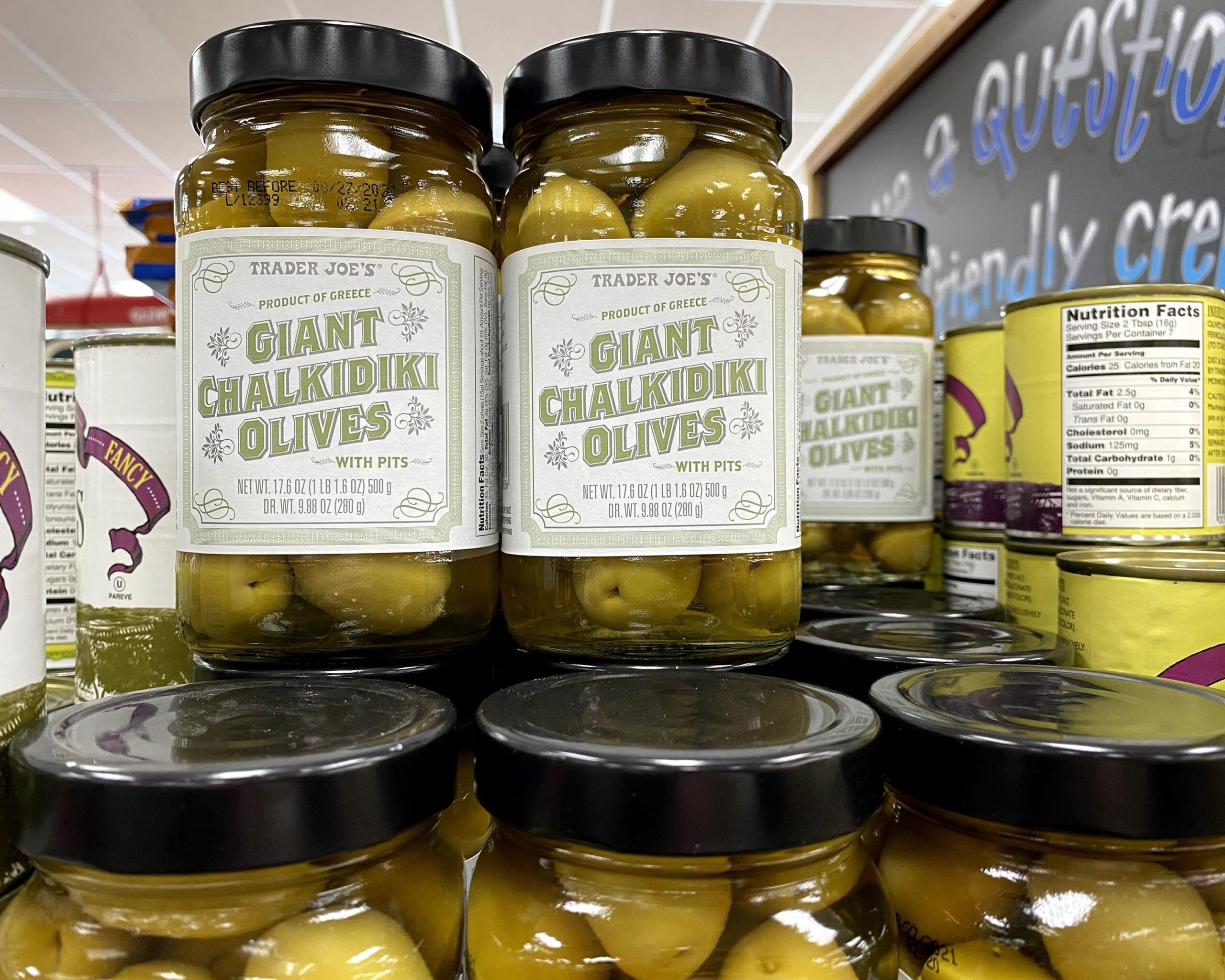 Three rows of jars of Trader Joe's Giant Chalkidiki Olives at a Trader Joe's, other rows of cans on both sides, white ceiling blurred in the background