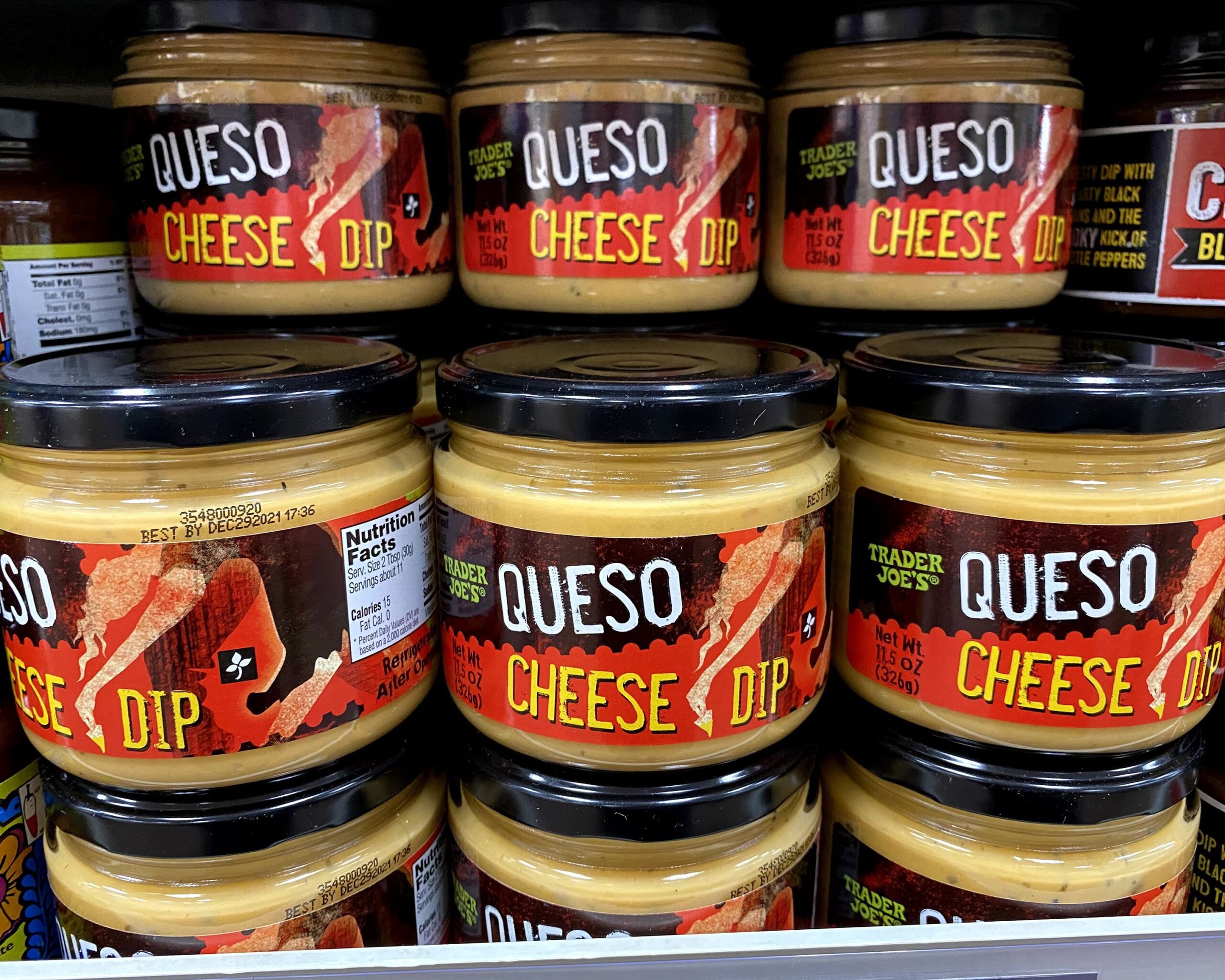 Three rows of jars of Trader Joe's Queso Cheese Dip at a Trader Joe's, on a shelf, jars of other dips on both sides