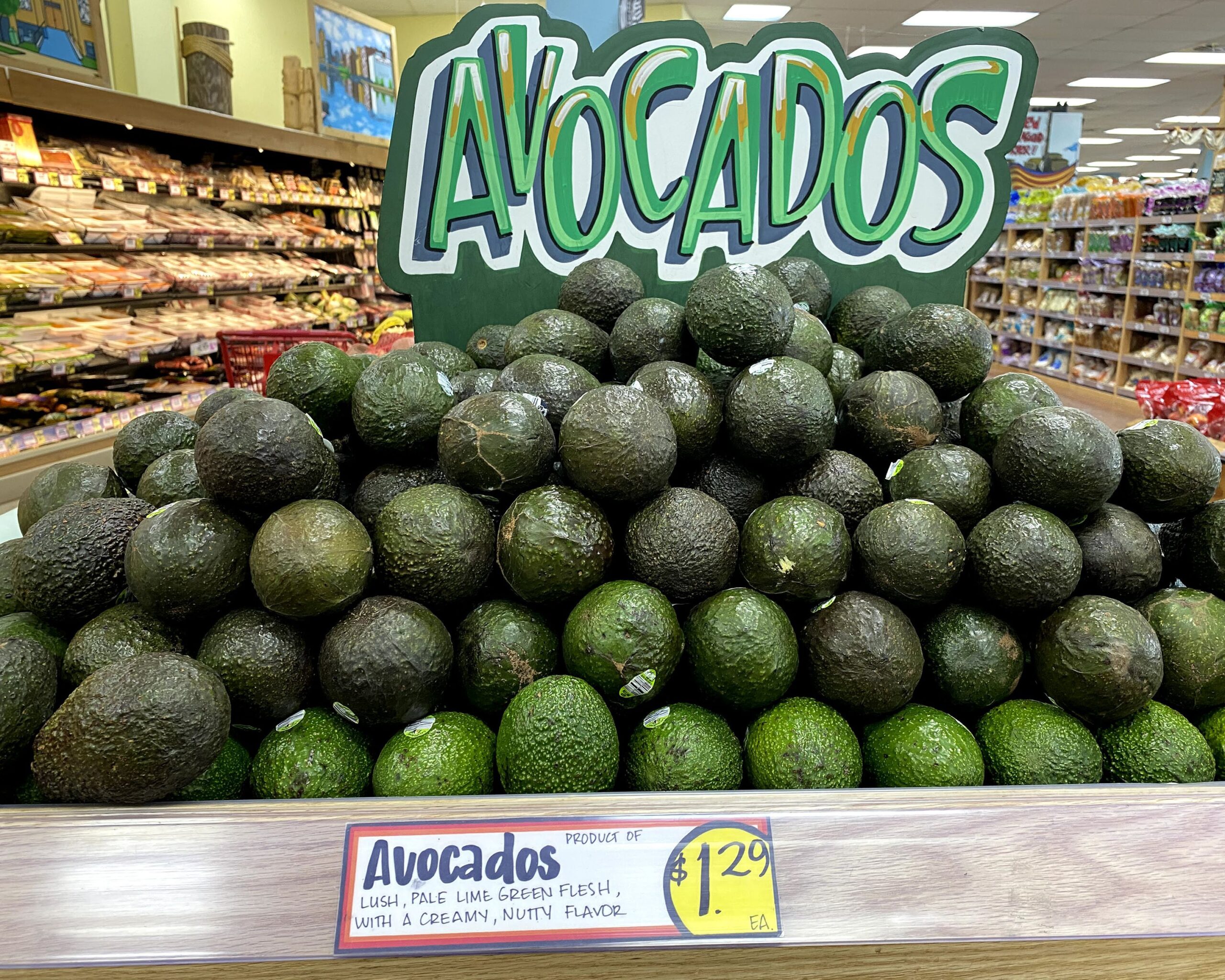 Large pile of Avocados at a Trader Joe's, fun large sign on top of it, in the produce section with meats on the left and breads on the right background