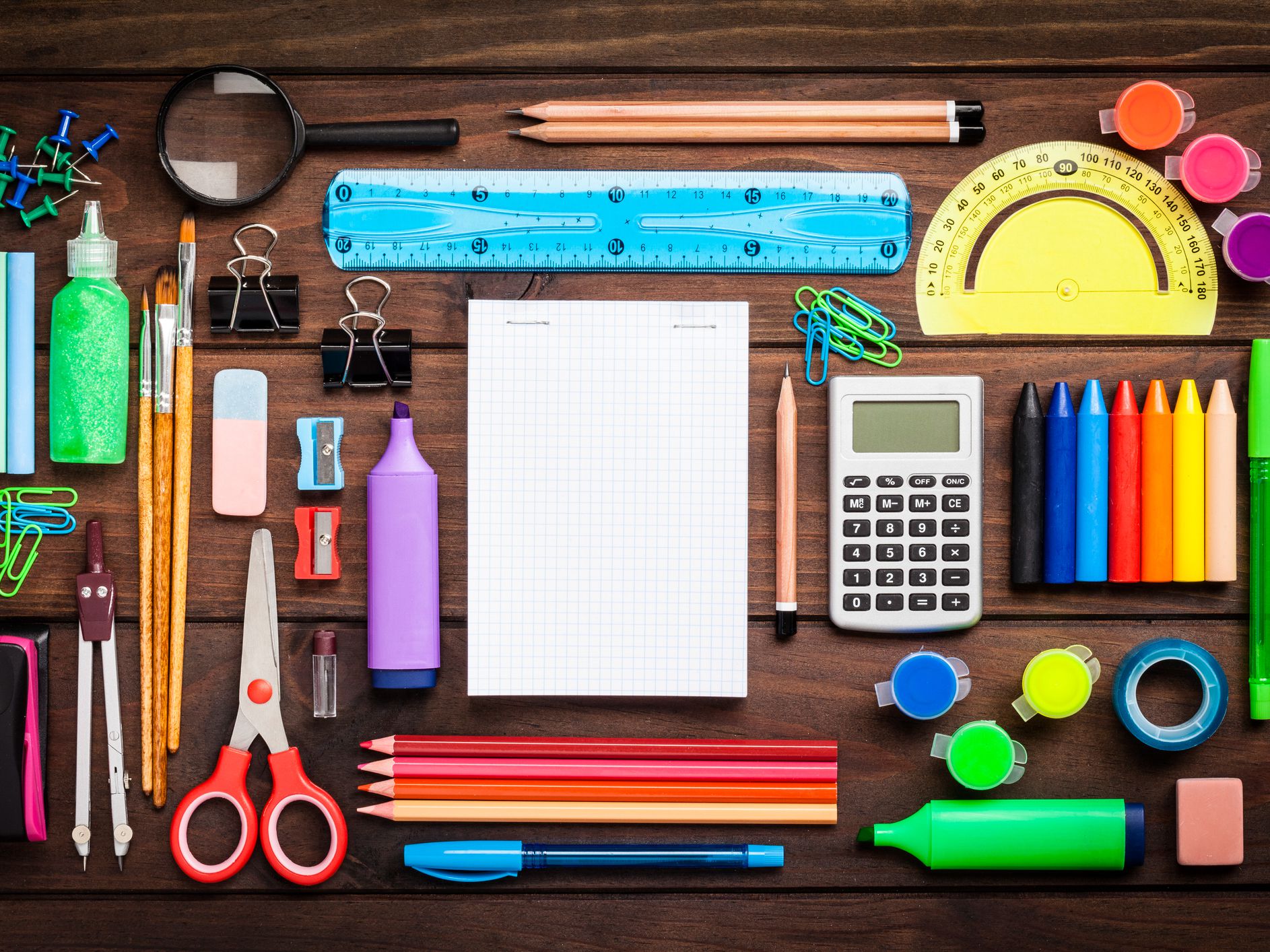 Top view of a large group of multi colored school or office supplies shot on rustic dark wooden table. A blank note pad is at the center of the composition with useful copy space ready for text and / or logo. DSRL studio photo taken with Canon EOS 5D Mk I