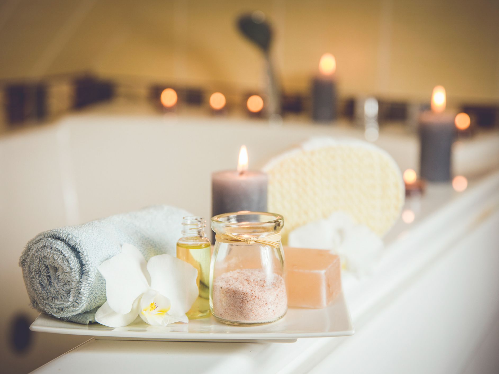 White ceramic tray with home spa supplies in home bathroom for relaxing rituals. Candlelight, salt soap bar, bath salt in jar, massage, bath oil in bottle, blue rolled towel, natural sponge.