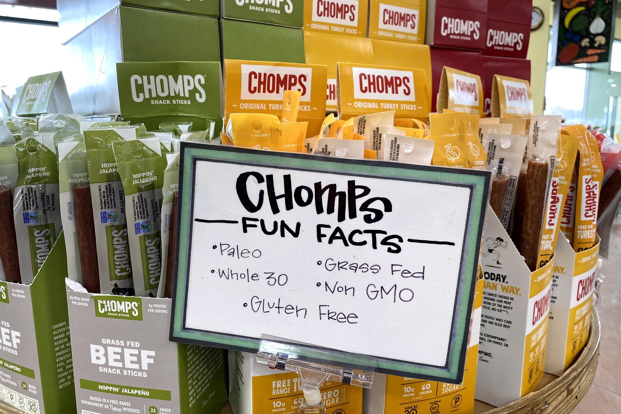 Packages of Chomps meat sticks at a Trader Joe's, boxes on top of one another in a circle, seating area and floor in the right background