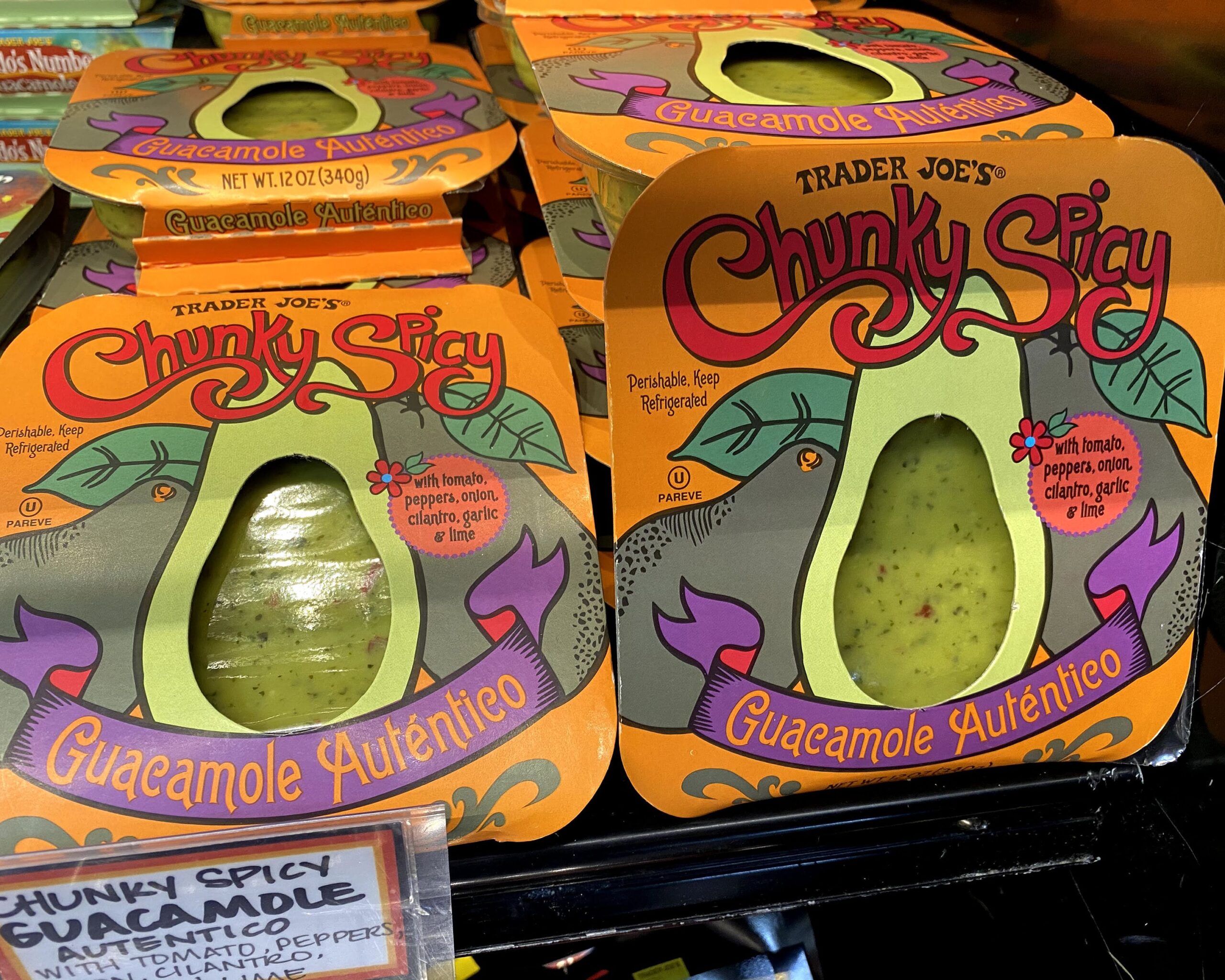 Two rows of Trader Joe's Chunky Spicy Guacamole packages at a Trader Joe's, in a refrigerator on a shelf, fun sign in the left foreground