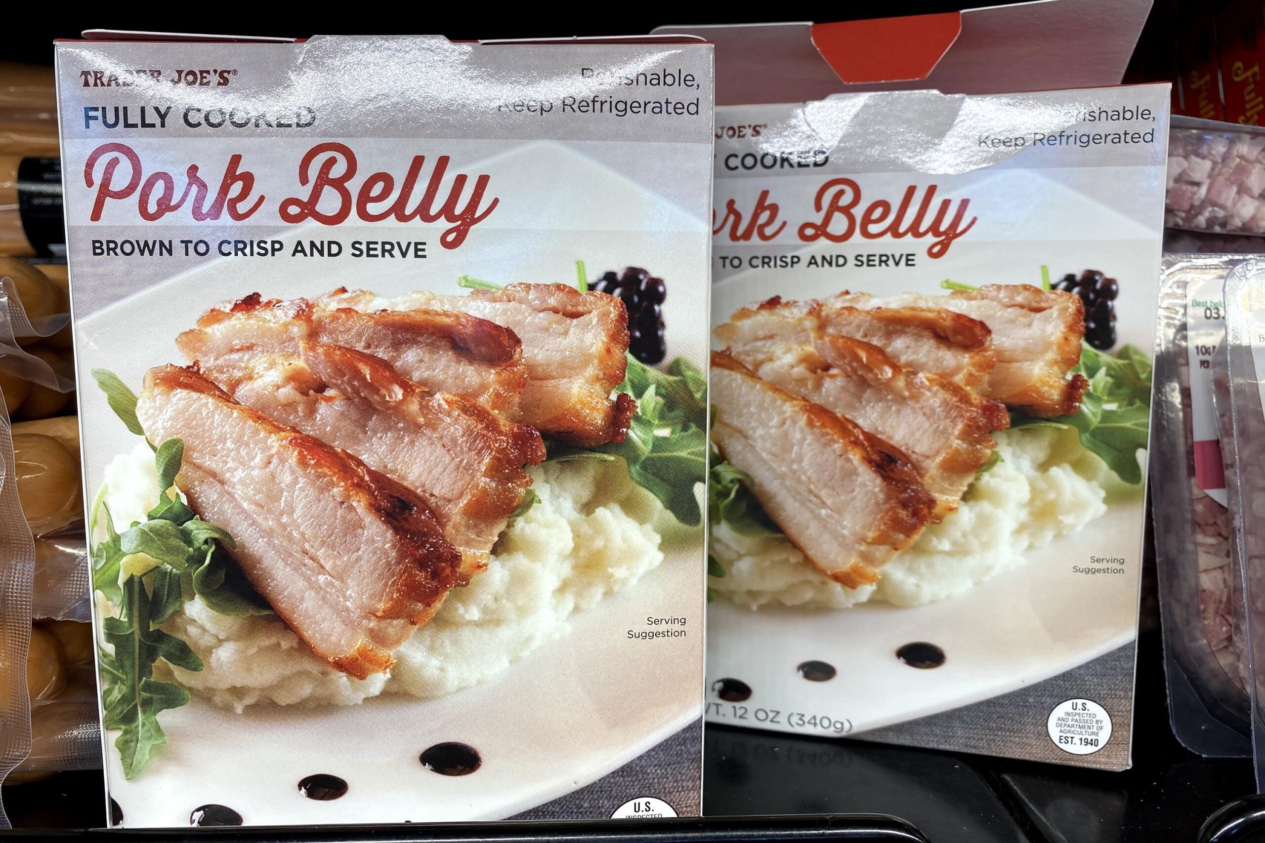 Two Trader Joe's Fully Cooked Pork Belly packages at a Trader Joe's, in a refrigerator on a shelf, other items for sale on both sides