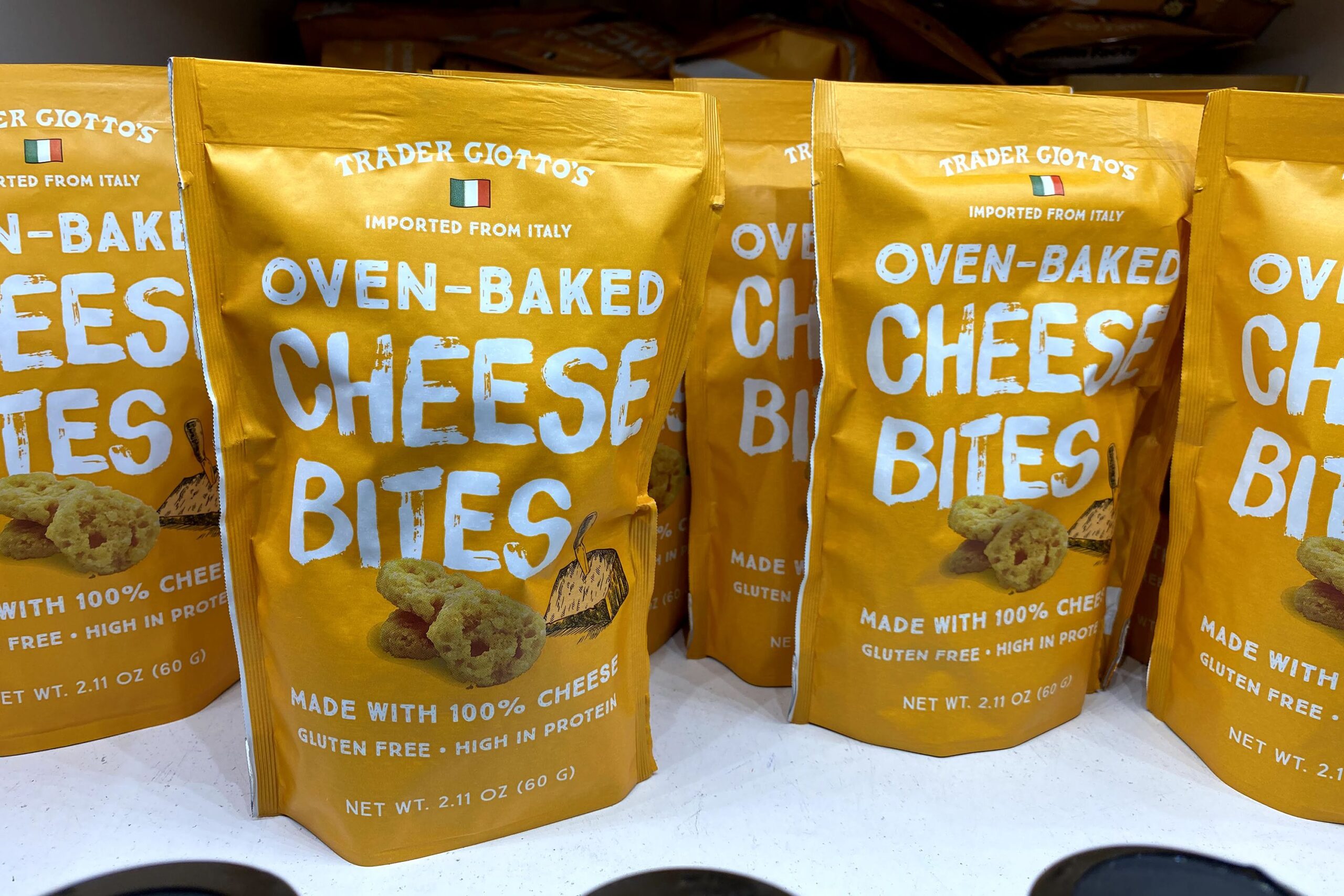 Many packages of Trader Joe's Oven Baked Cheese Bites at a Trader Joe's, on a white cardboard shelf