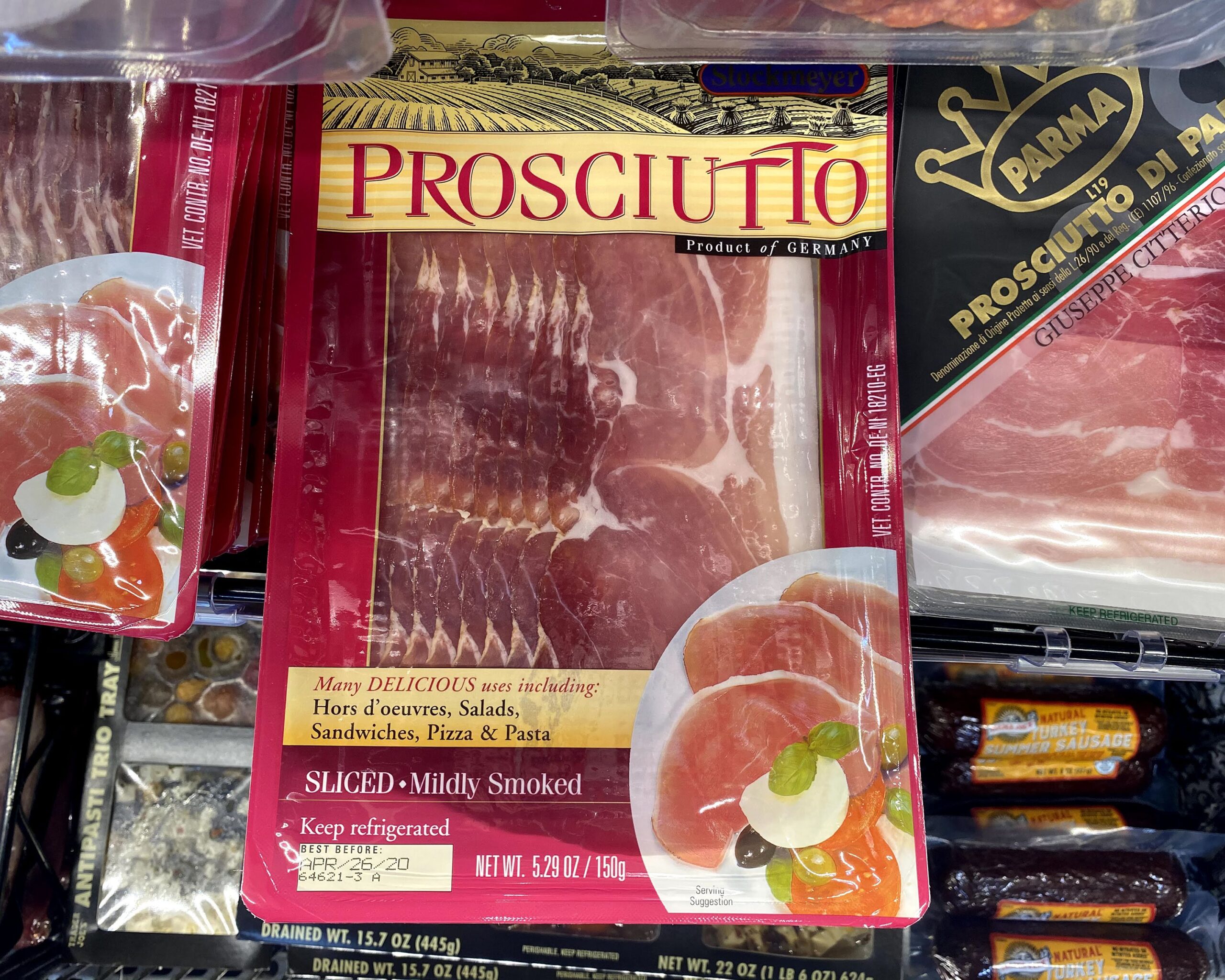 Row of of Trader Joe's Prosciutto packages at a Trader Joe's in a refrigerator, surrounded by other speciality meats, antipasti, and olives