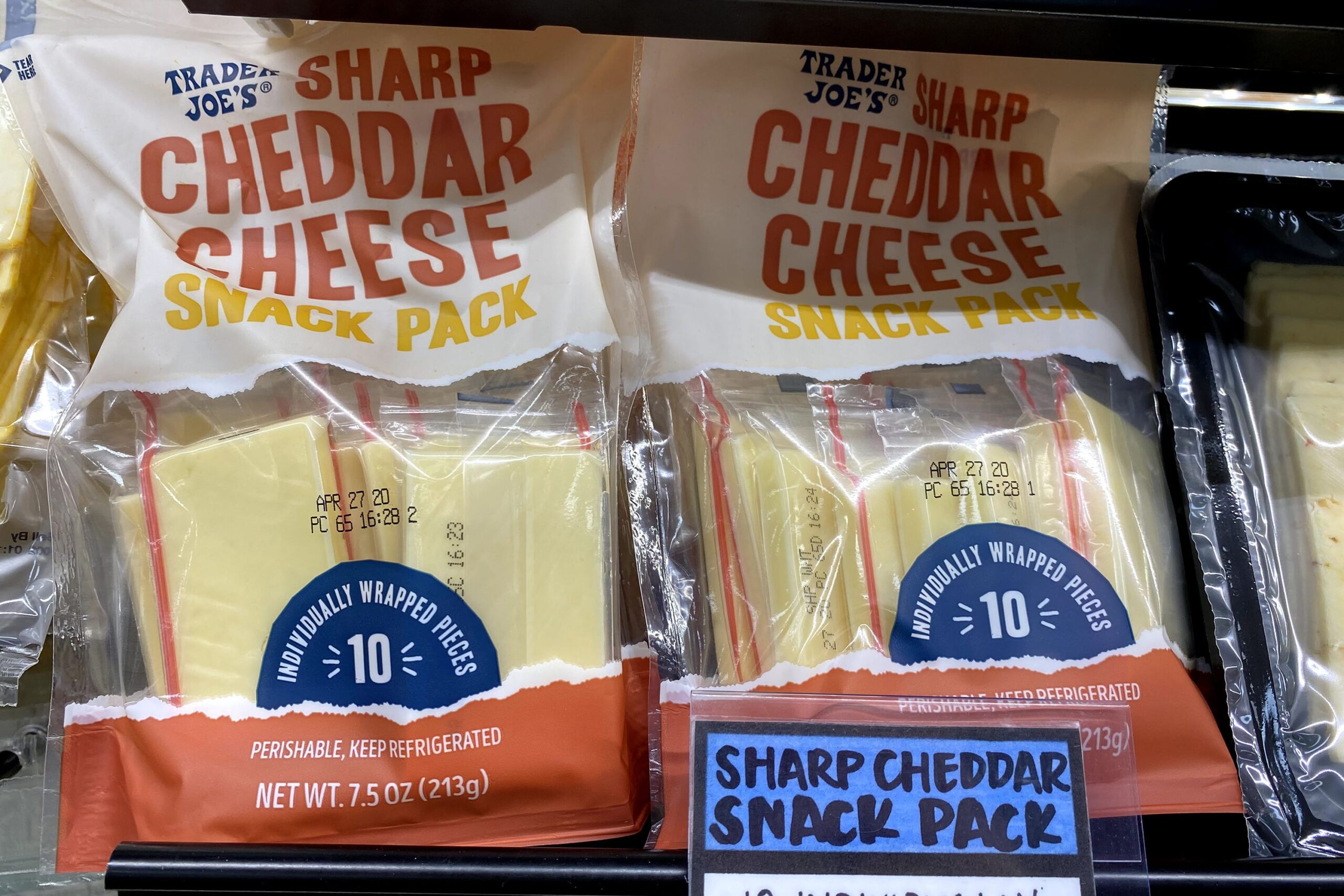 Two rows of packages of Trader Joe's Sharp Cheddar Snack Pack at a Trader Joe's in a refrigerator on a shelf, other packages of cheese on both sides