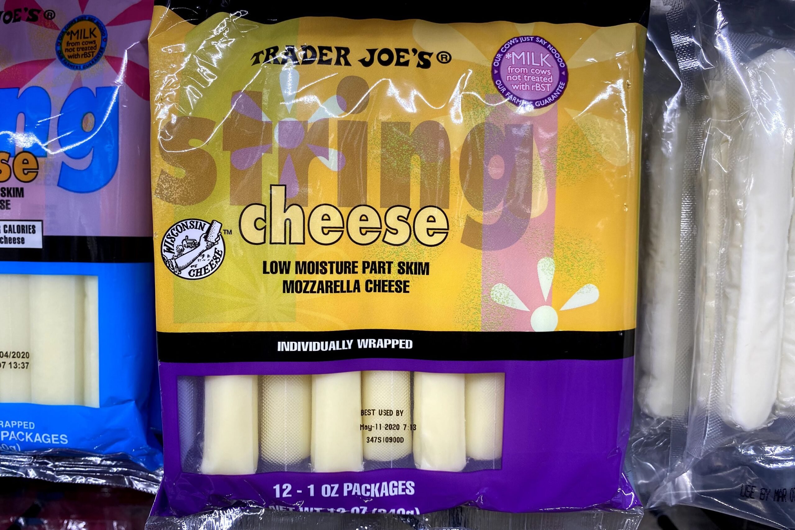A row of packages of Trader Joe's String Cheese at a Trader Joe's, in a refrigerator, two other packages of string cheeses on both sides