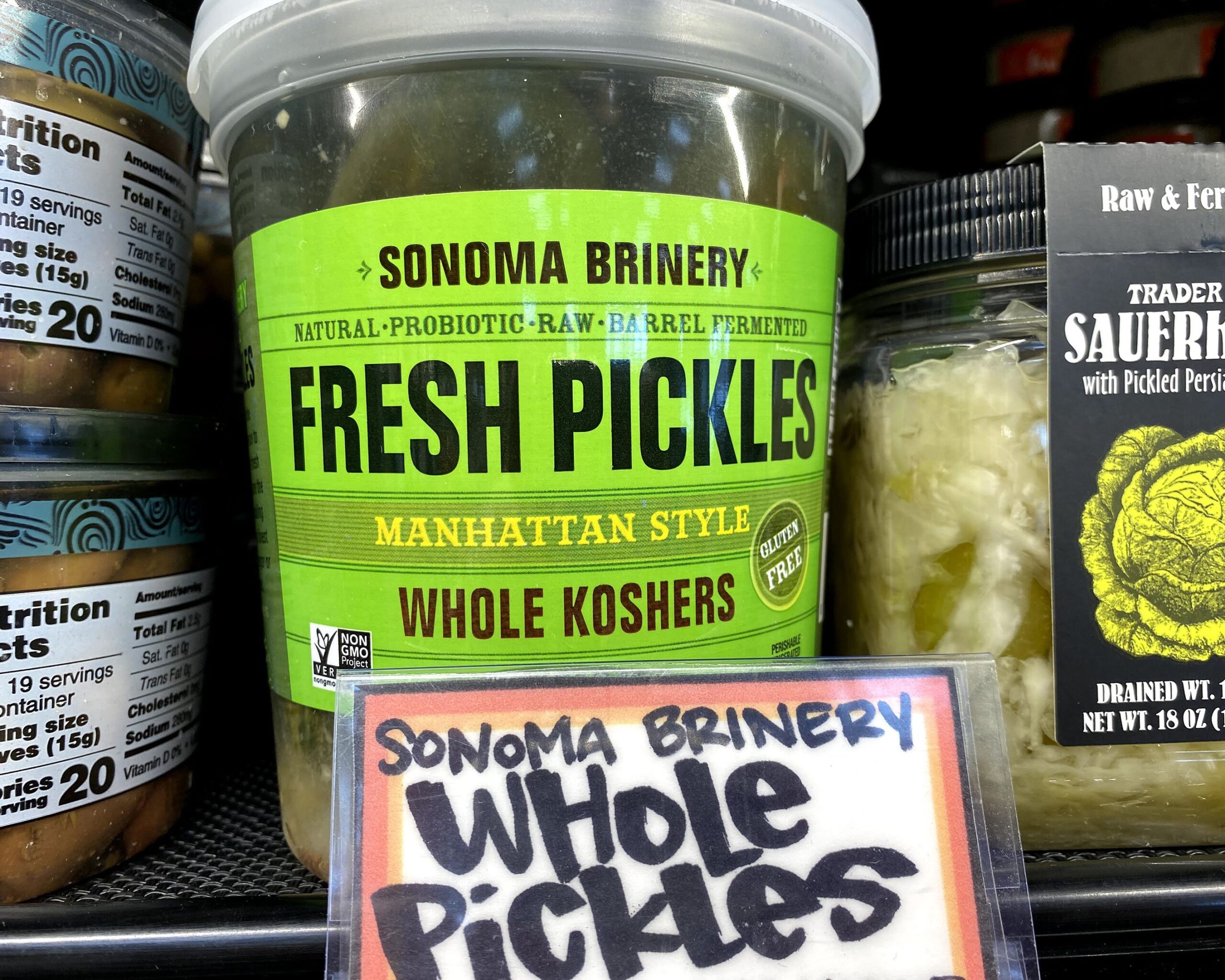 Plastic jar of Sonoma Brinery Fresh Pickles at a Trader Joe's, in a refrigerator on a shelf, jars of olives on the left and jars of sauerkraut on the right, fun sign in the foreground