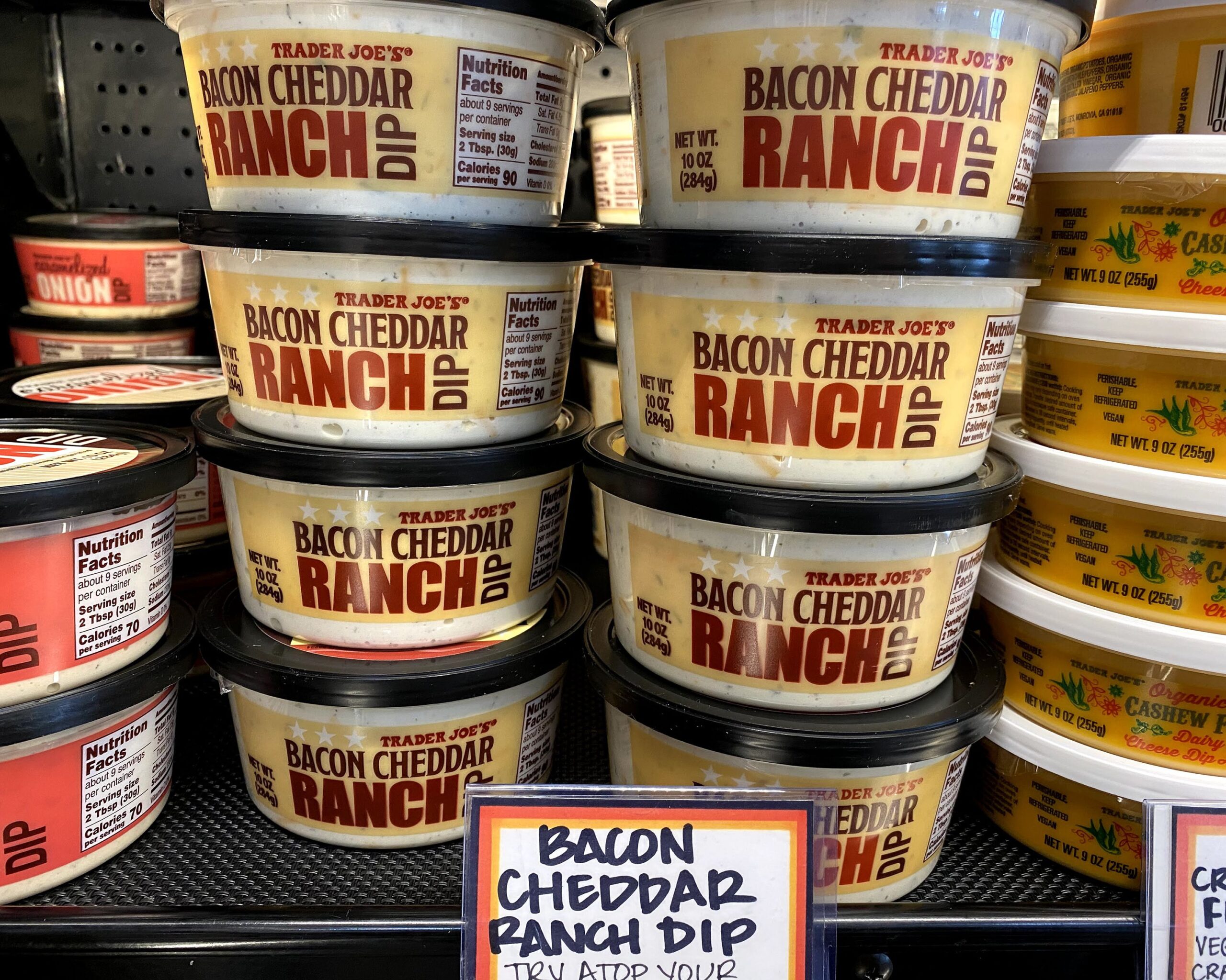 Two rows of tubs of Trader Joe's Bacon Cheddar Ranch Dip at a Trader Joe's, in a refrigerator on a shelf, other tubs of dip on both sides and in the background