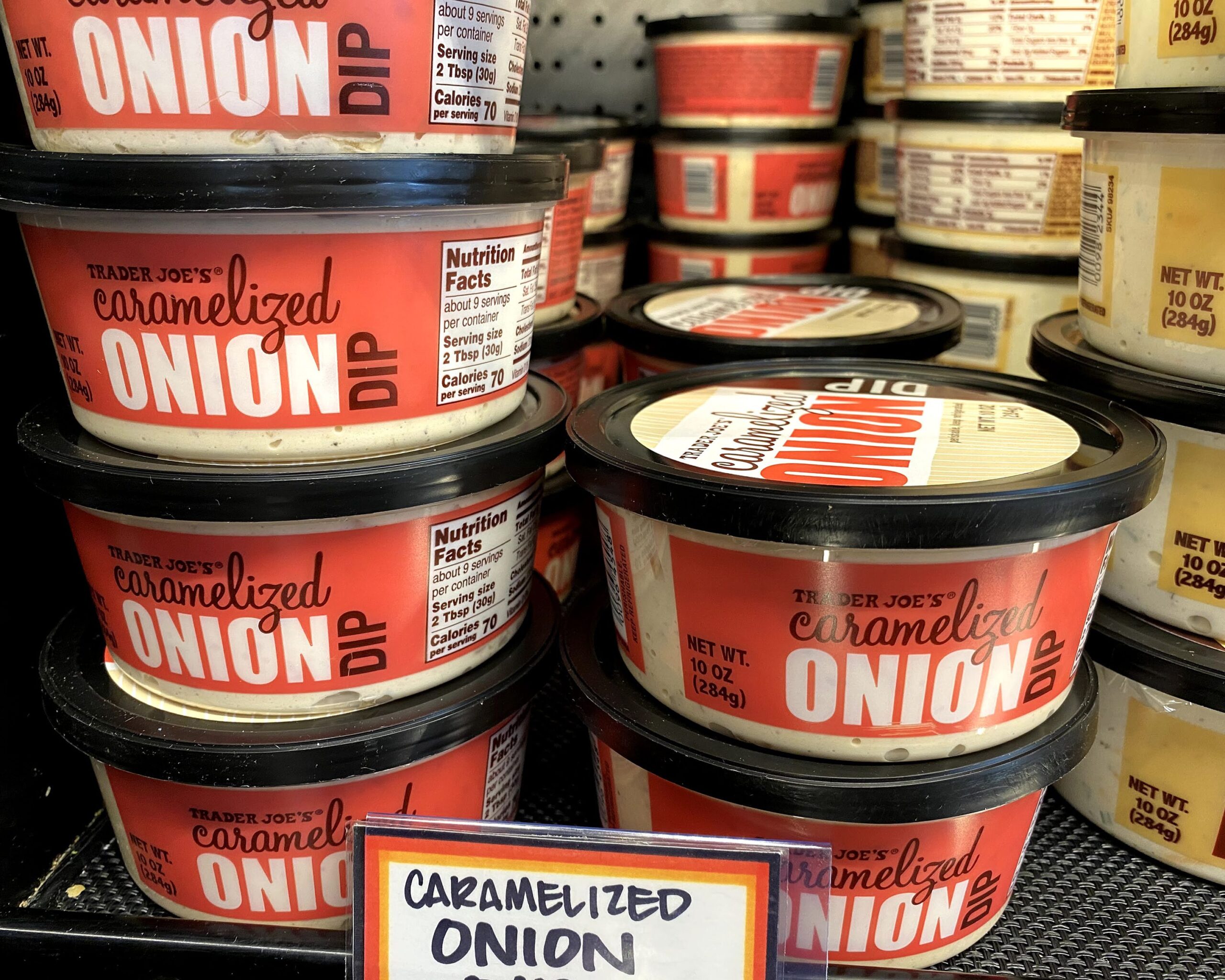 Two rows of tubs of Trader Joe's Caramelized Onion Dip at a Trader Joe's, on a shelf, several rows of tubs in the background, another kind on the right side