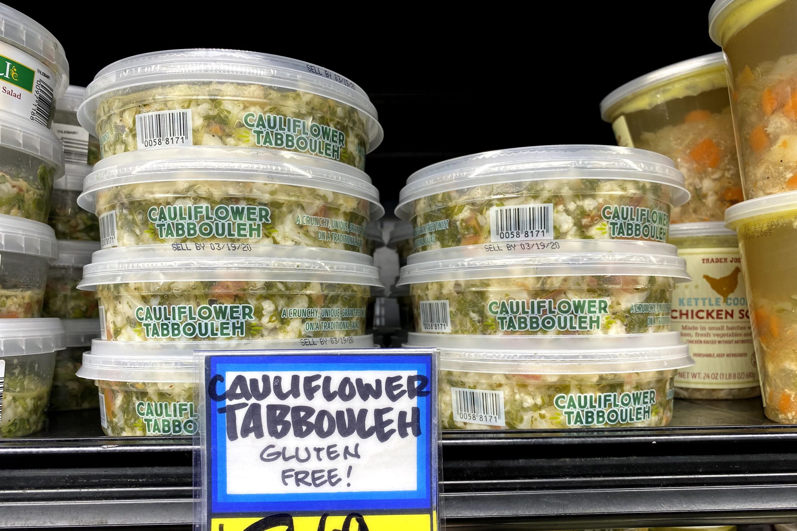 Two rows of tubs of Trader Joe's Cauliflower Tabbouleh at a Trader Joe's, in a refrigerator on a shelf, tubs of sauce on the left side and tubs of a soup on the right