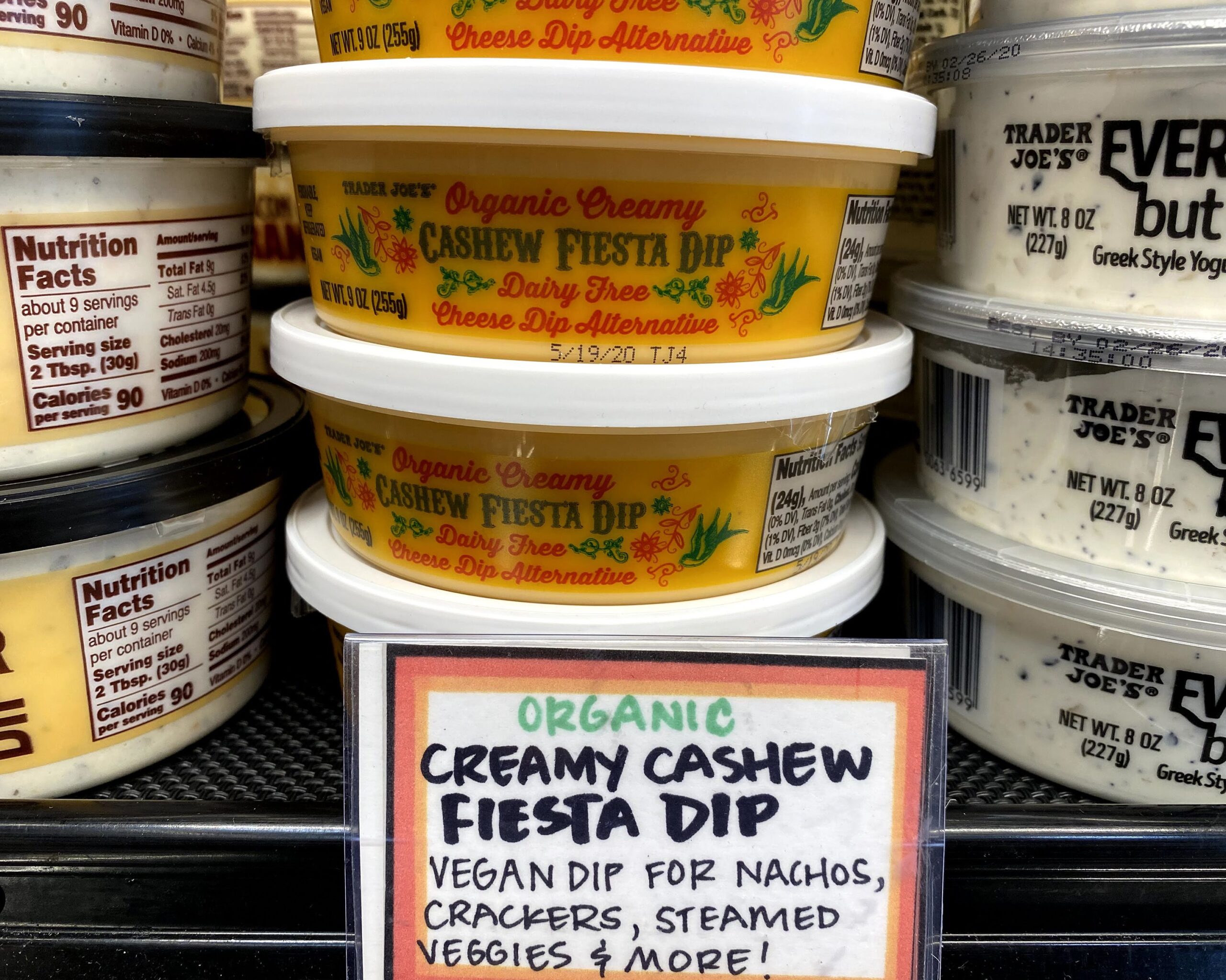 Tubs of Trader Joe's Creamy Cashew Fiesta Dip at a Trader Joe's, piled on each other, in a refrigerator, tubs of other dips on both sides