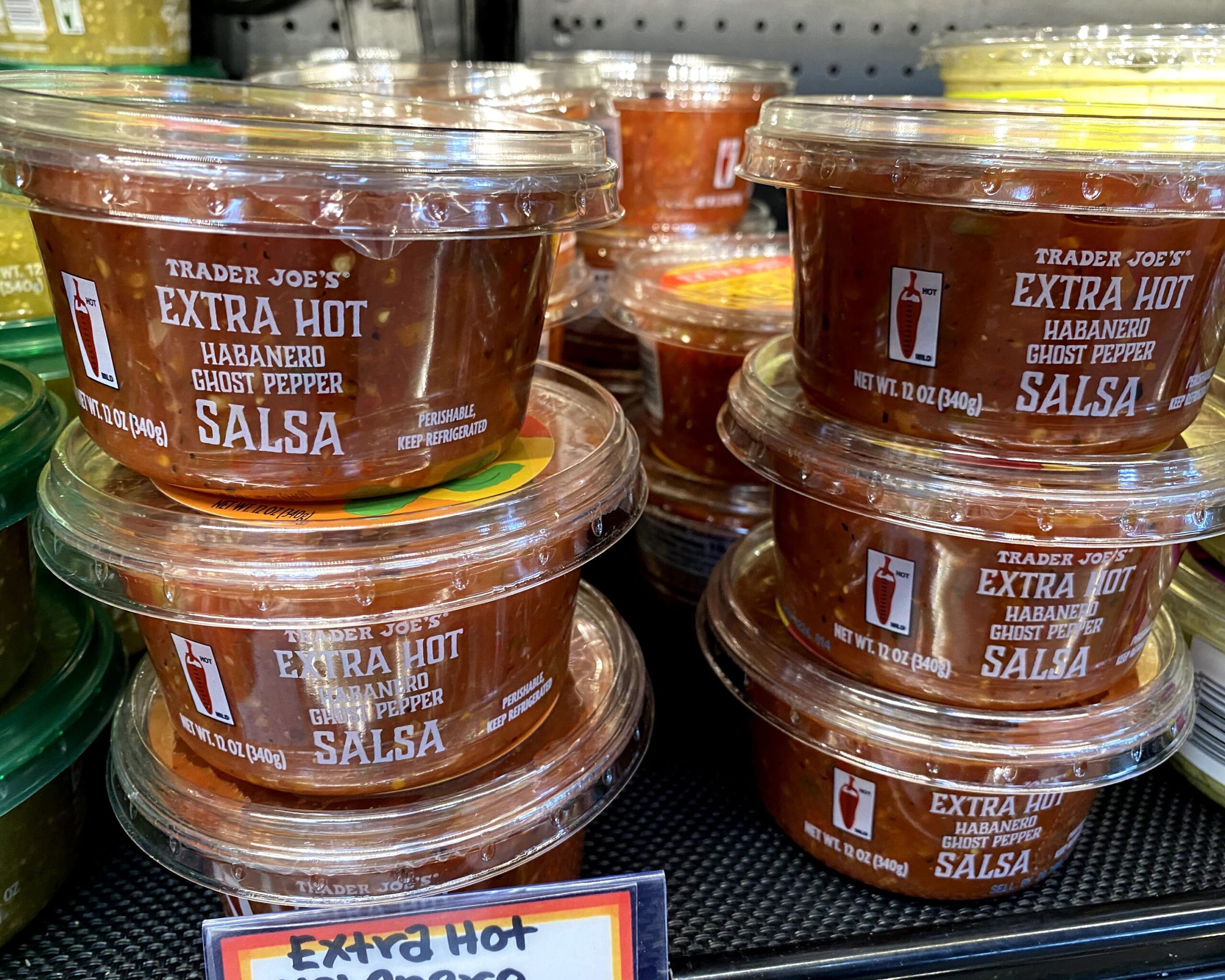 Many stacks of tubs of Trader Joe's Extra Hot Habanero Ghost Pepper Salsa at a Trader Joe's, in a refrigerator on a shelf, other tubs of dips on both sides
