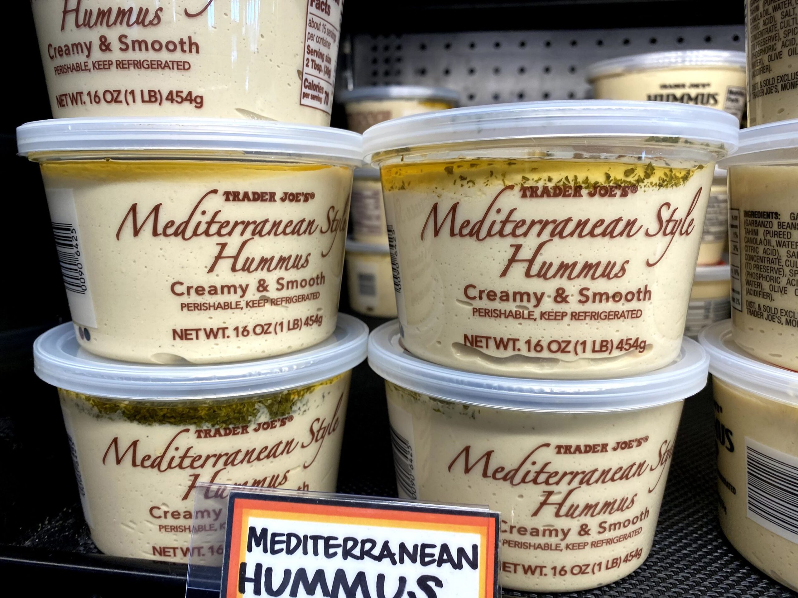 Many Tubs of Trader Joe's Mediterranean Style Hummus at a Trader Joe's in the refrigerator, more hummus tubs in the background