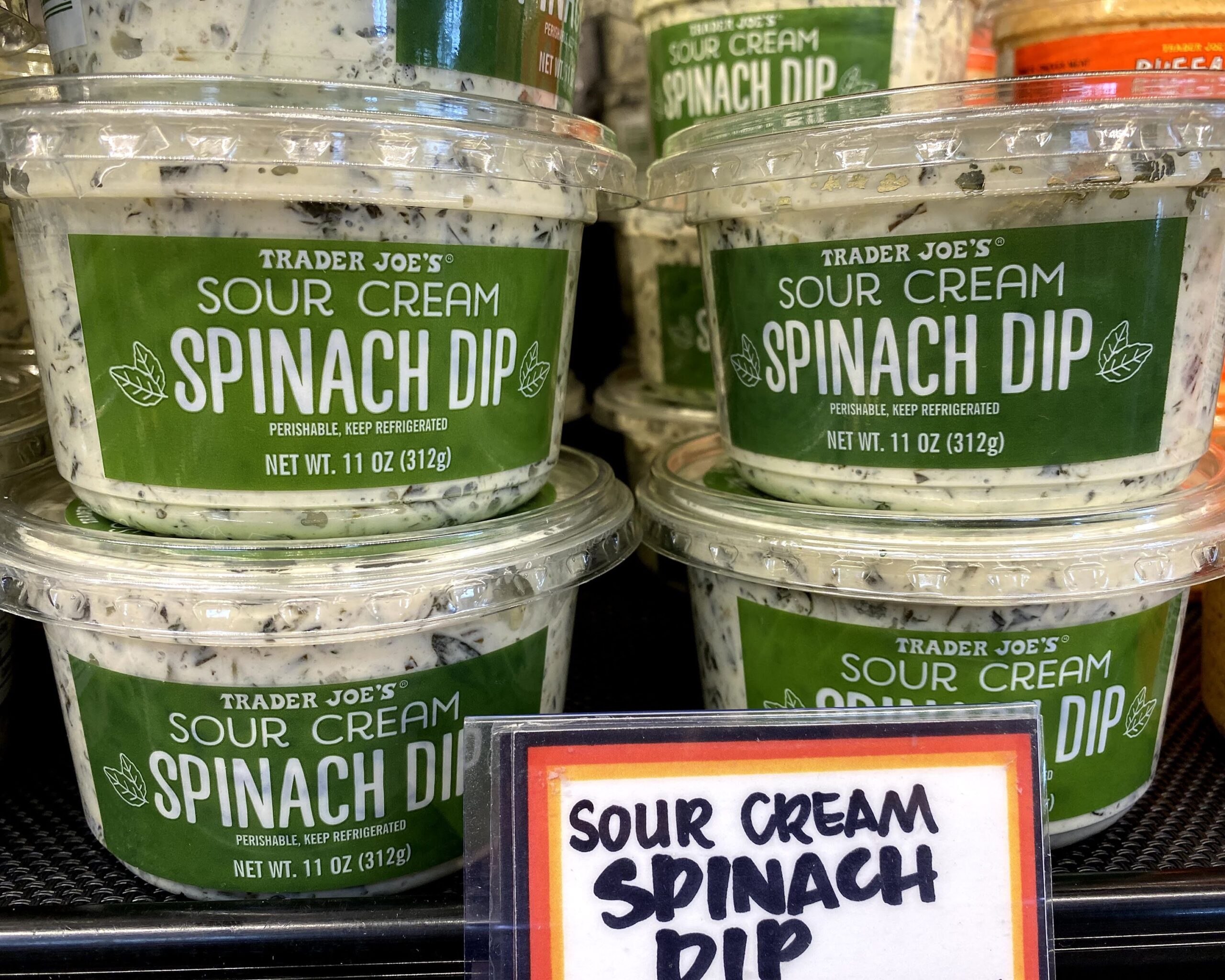 Two rows of tubs of Trader Joe's Spinach Dip at a Trader Joe's in a refrigerator on a shelf, several tubs in the background