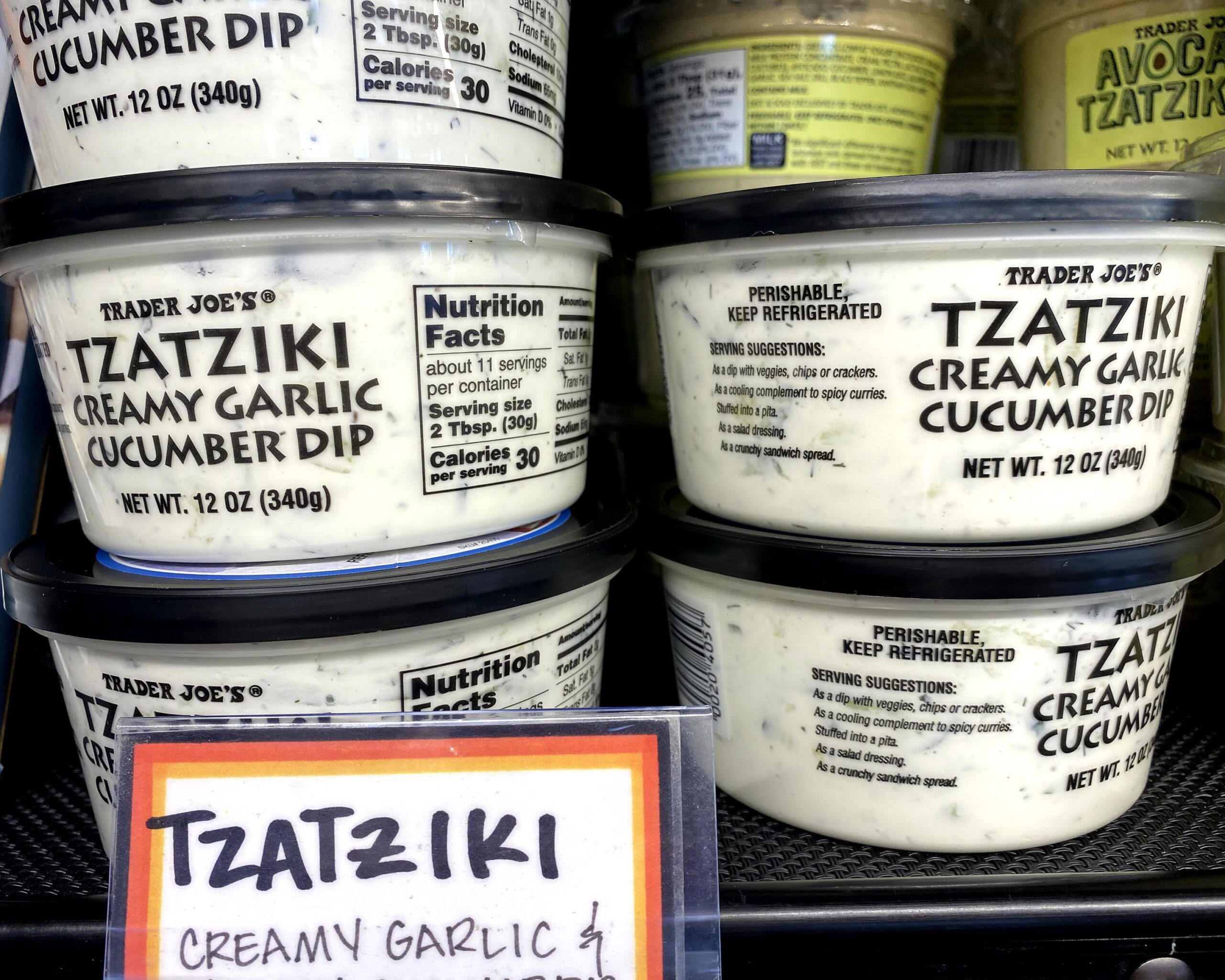 Two rows of tubs of Trader Joe's Tzatziki at a Trader Joe's, in a refrigerator on a shelf, fun sign in the foreground, tubs of Trader Joe's Avocado Tzatziki in the right background