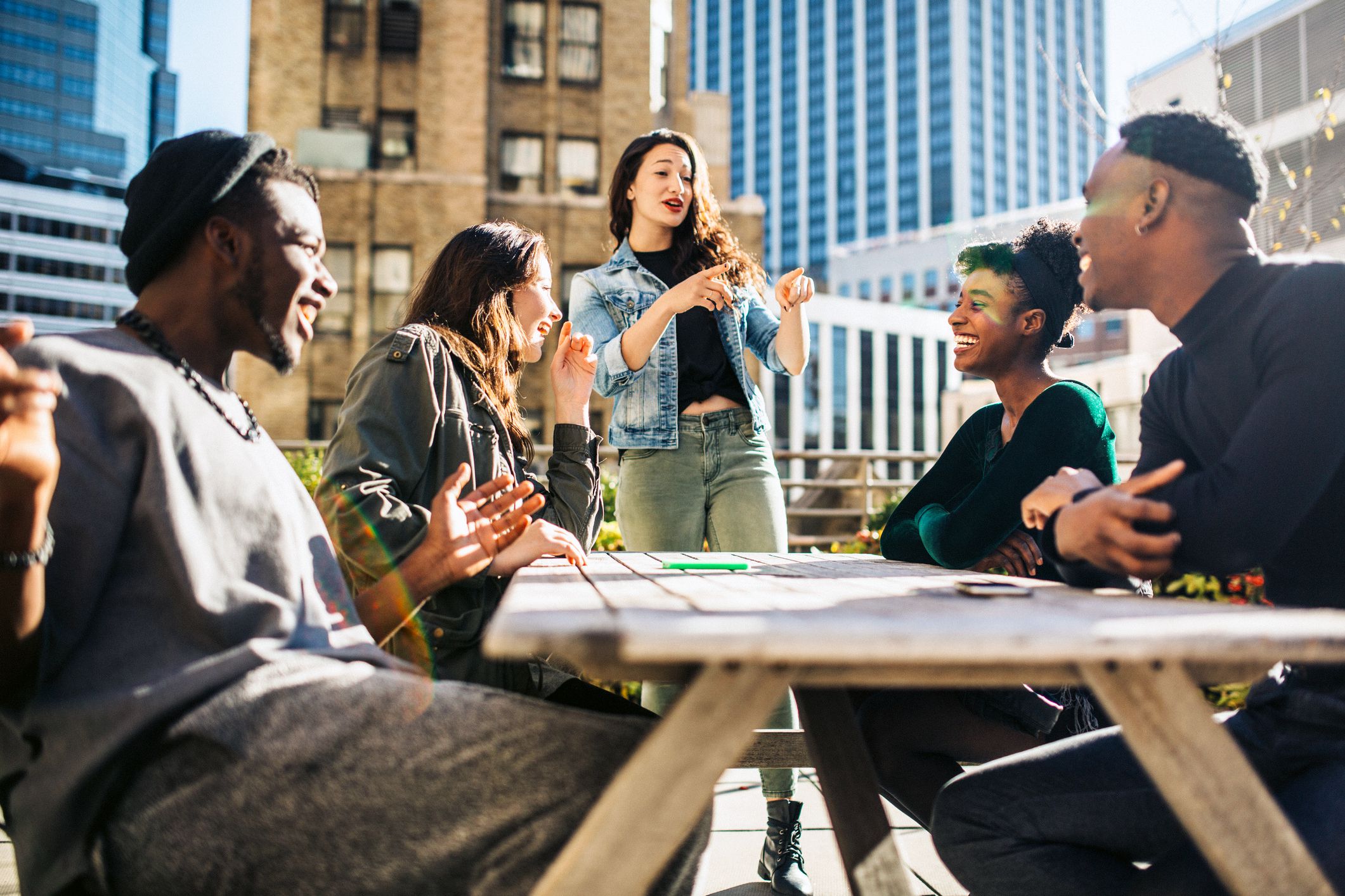 A group of friends smile and laugh while sitting at a wooden picnic table on a New York City rooftop
