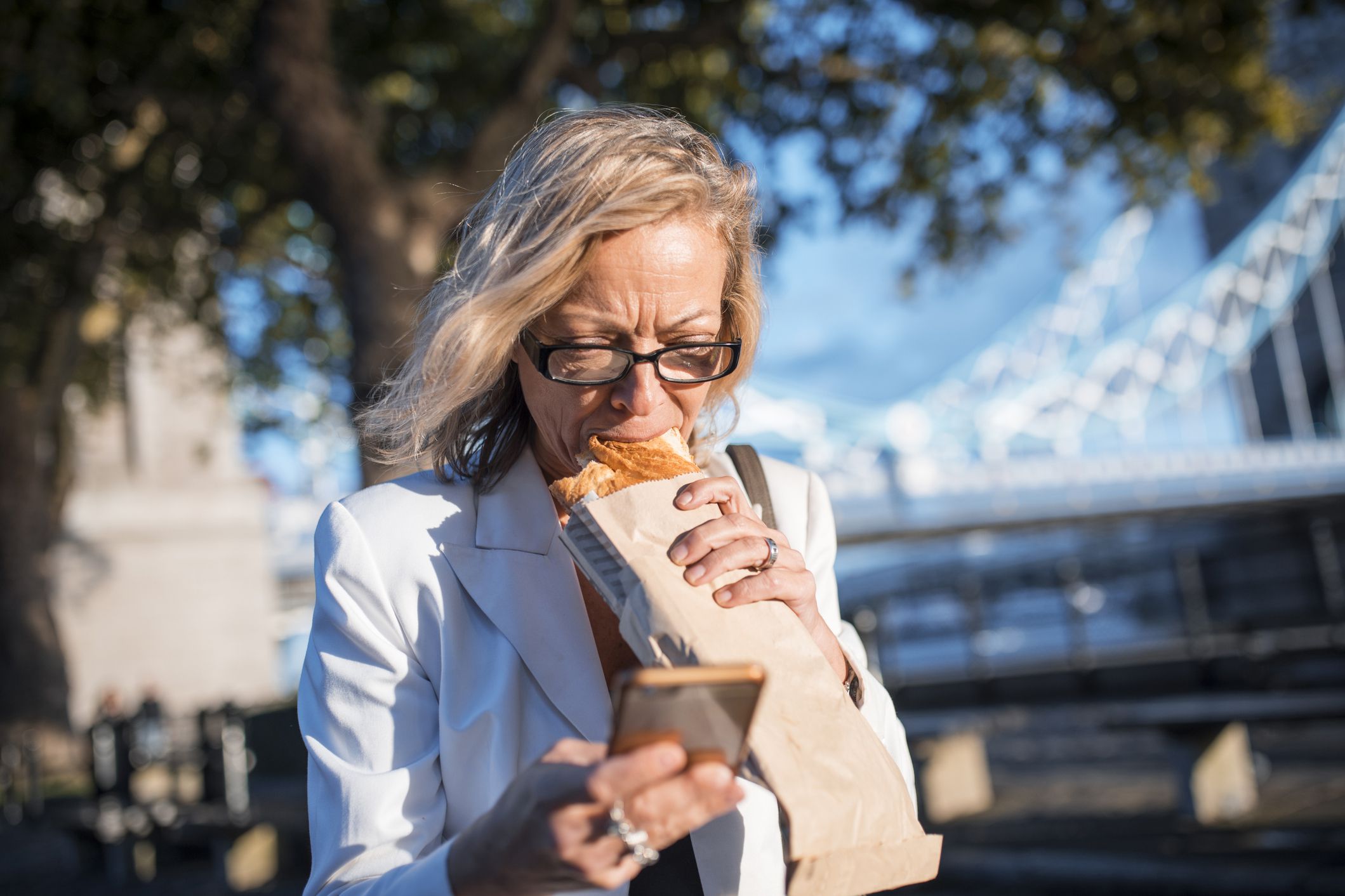 Mid adult businesswoman on the go. She is eating, using mobile phone and looking at it. Tower bridge in the back.