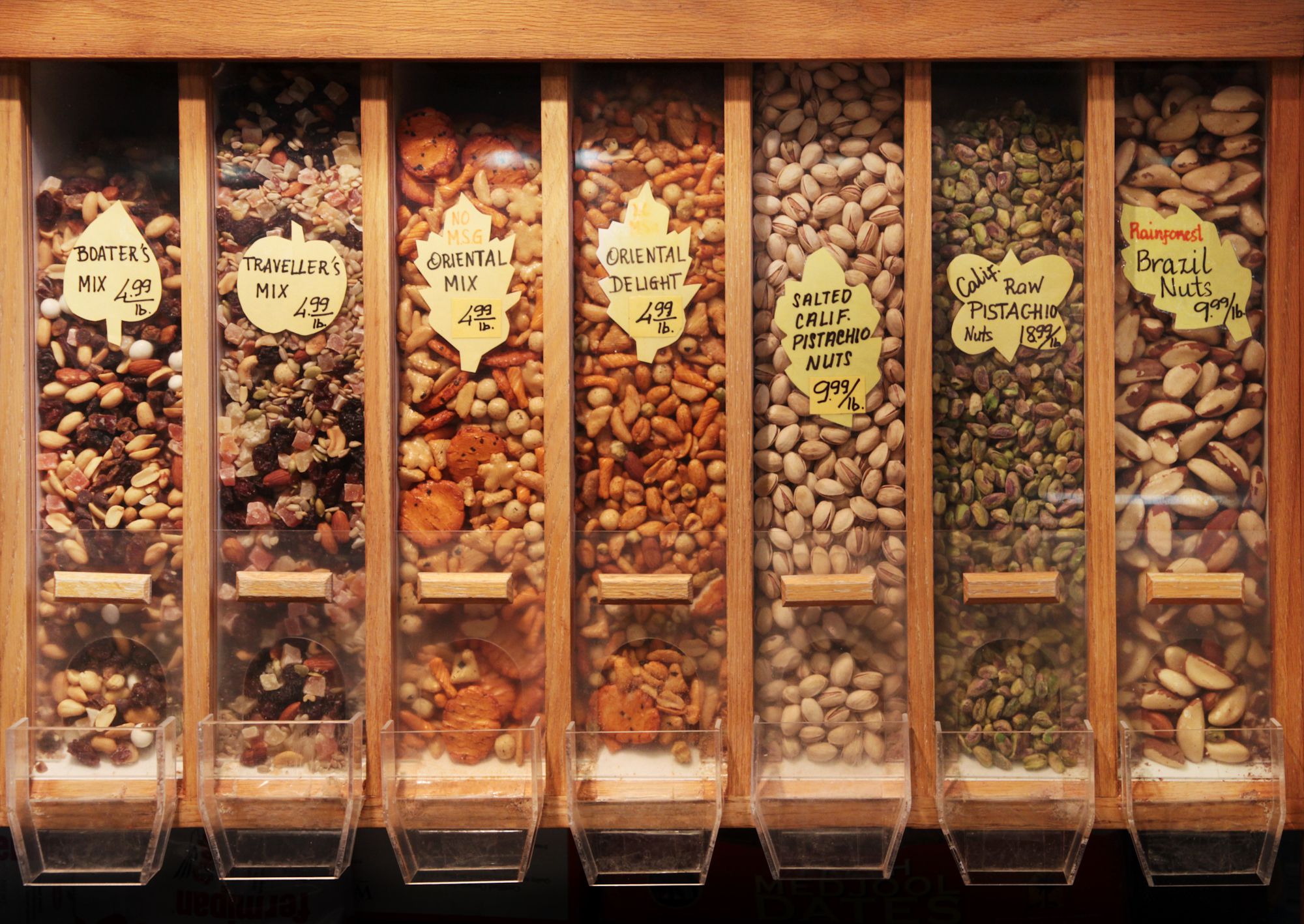 Seven types of trailmix and nuts in a dispenser at a public market.