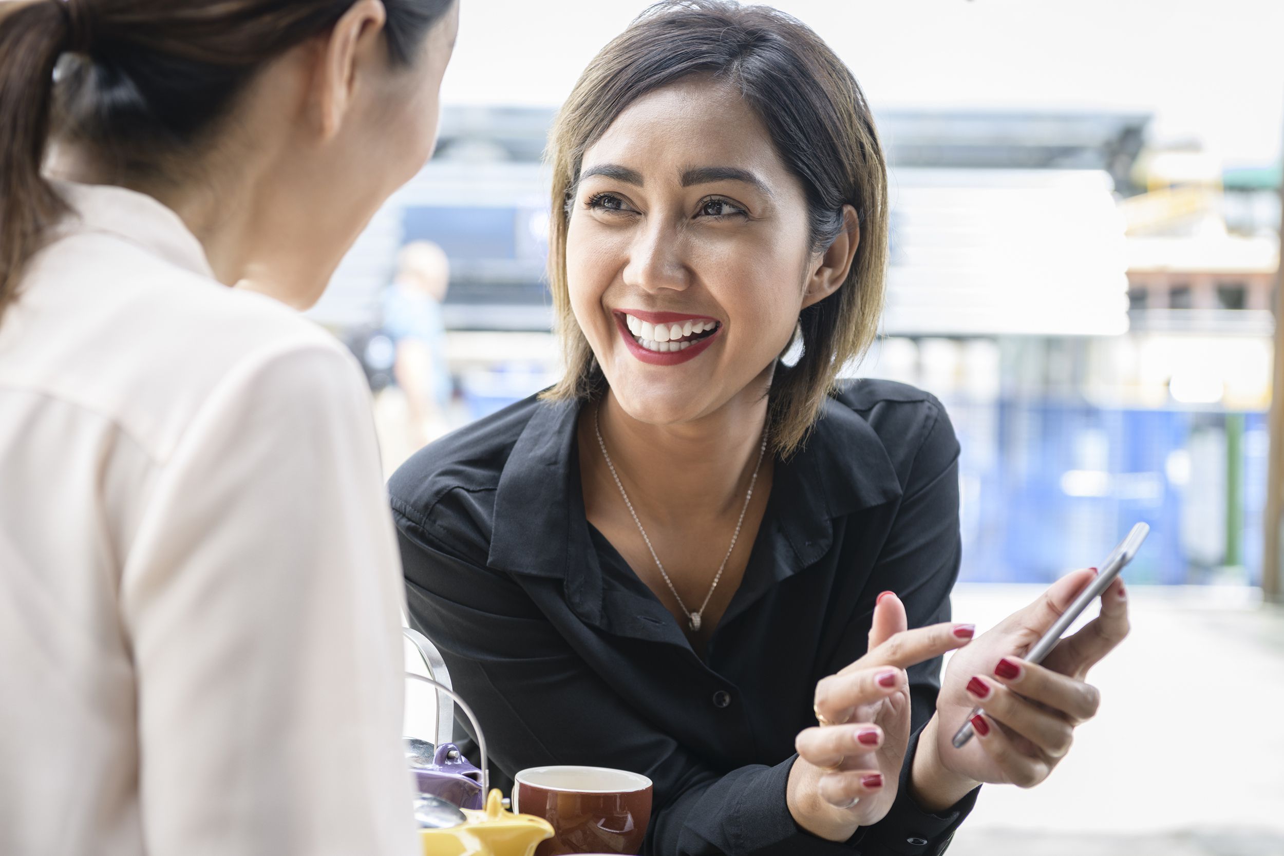 smiling asian woman showing friend social media on smartphone