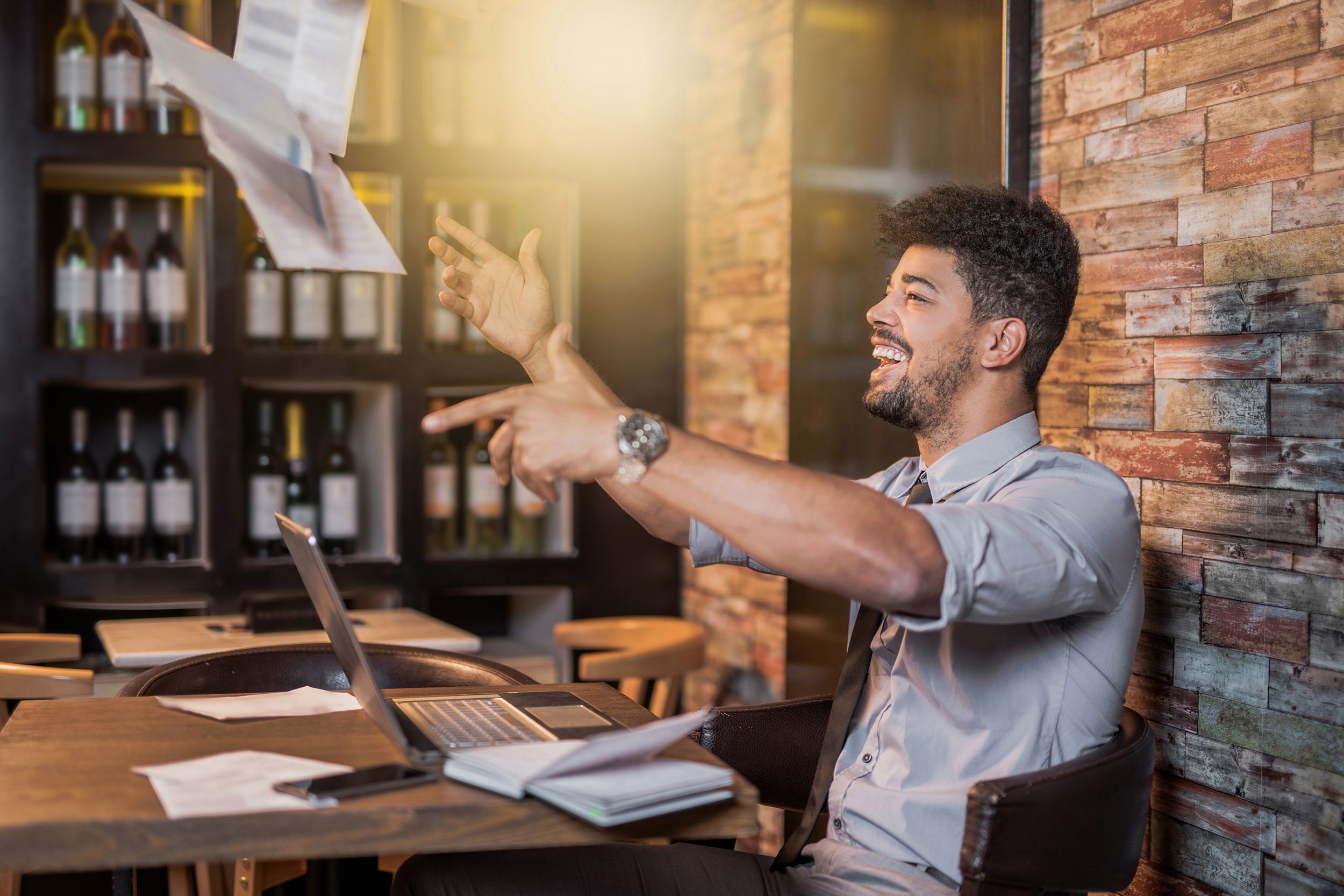 laughing man throwing papers in air while working on laptop in cafe