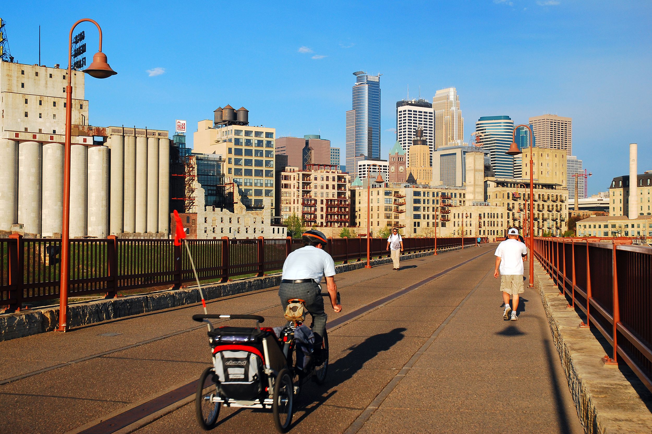 adult man commutes to work by bicycle, carrying his child in a trailer as he crosses the Stone Arch Bridge in Minneapolis