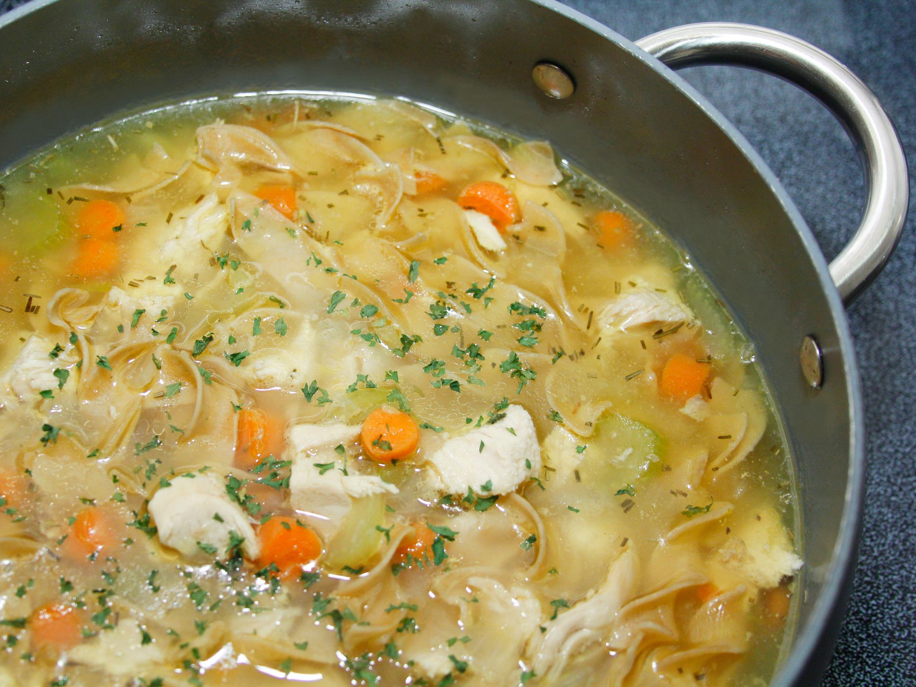 Homemade chicken noodle soup in pot with carrots, celery, topped with parsley bits.