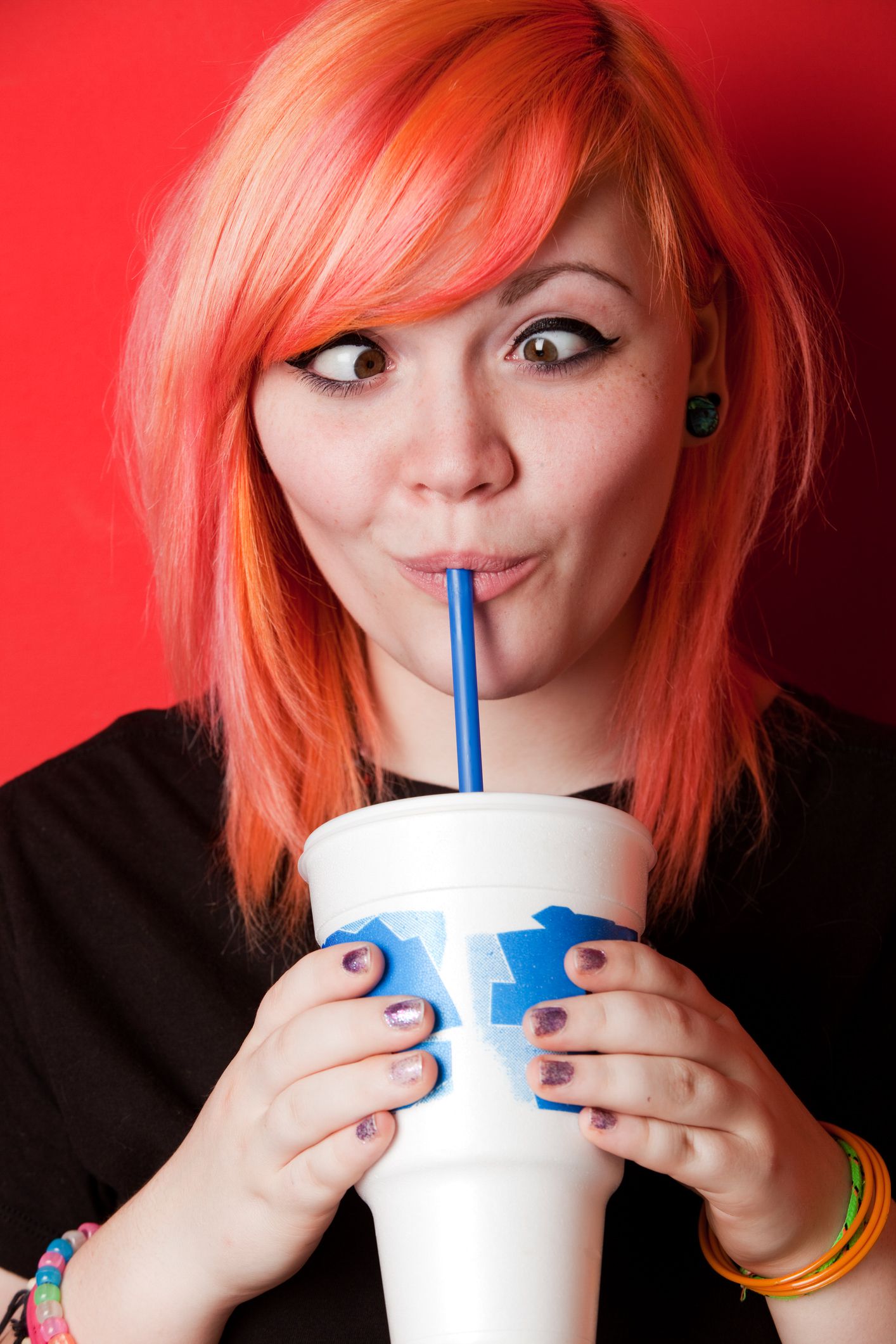Hip teen girl with hair dyed a peach color looks cross-eyed as she sucks soda from an extra large cup.