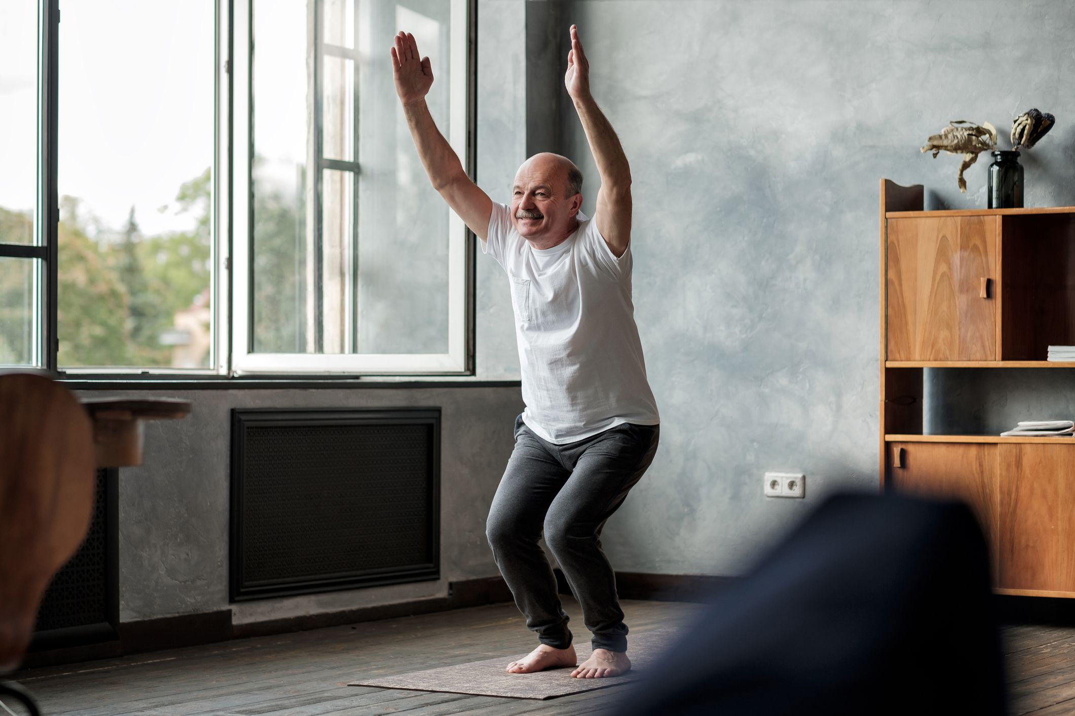 Senior man practicing yoga indoors at living room doing Chair pose or Utkatasana