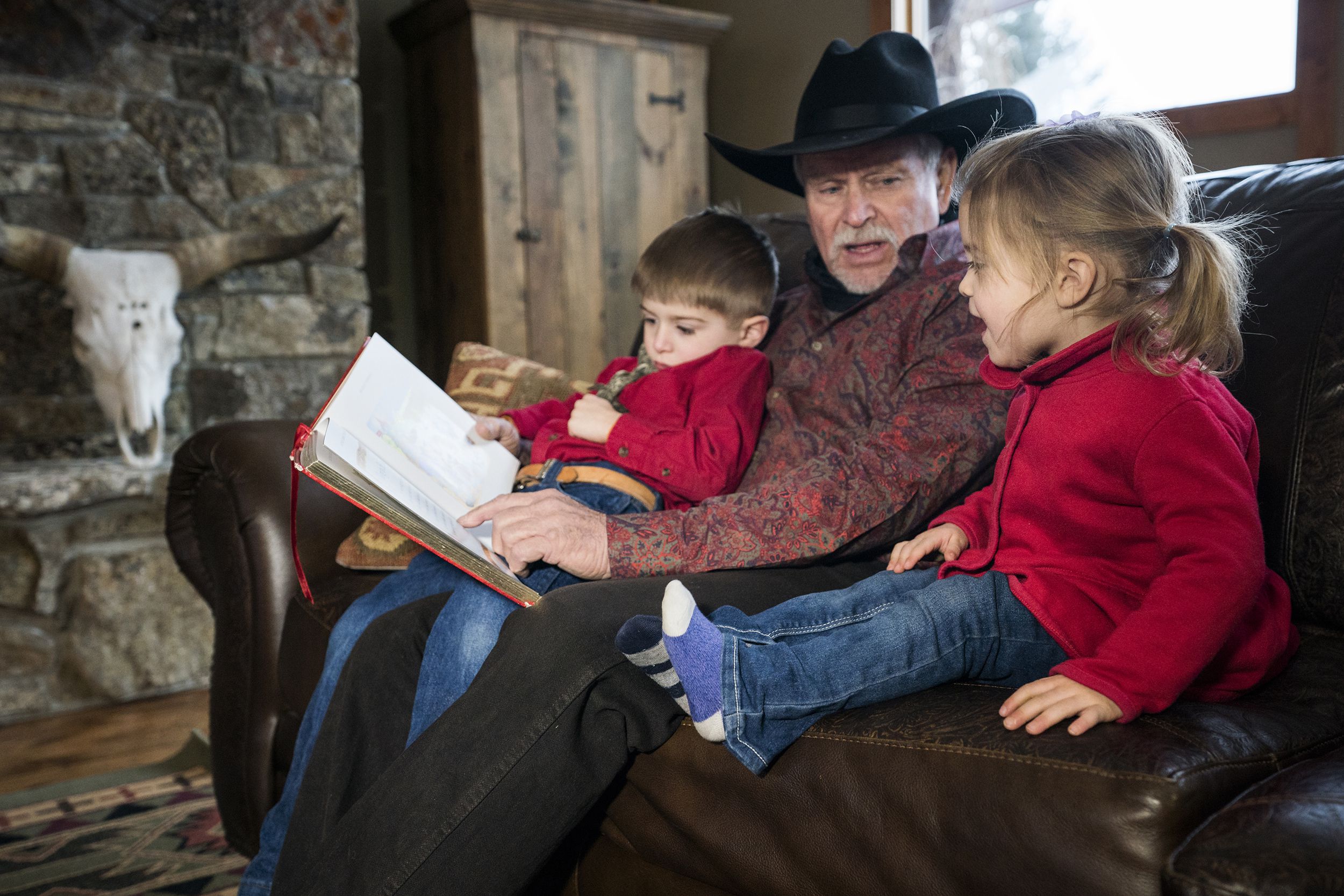 male cowboy reading a storybook to his yound grandchildren sitting in his lap, Montana