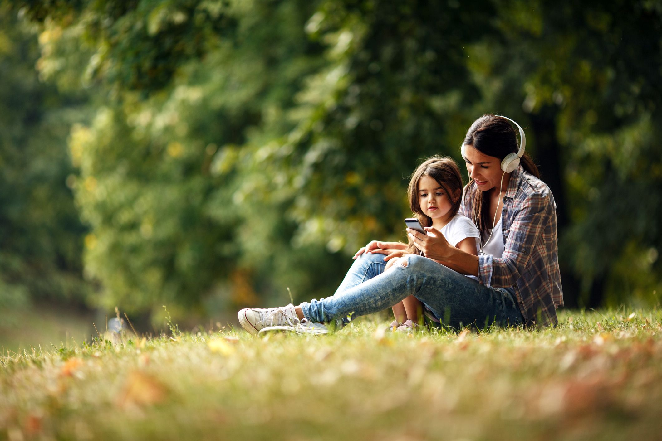 young woman and child looking at phone with headphones in park