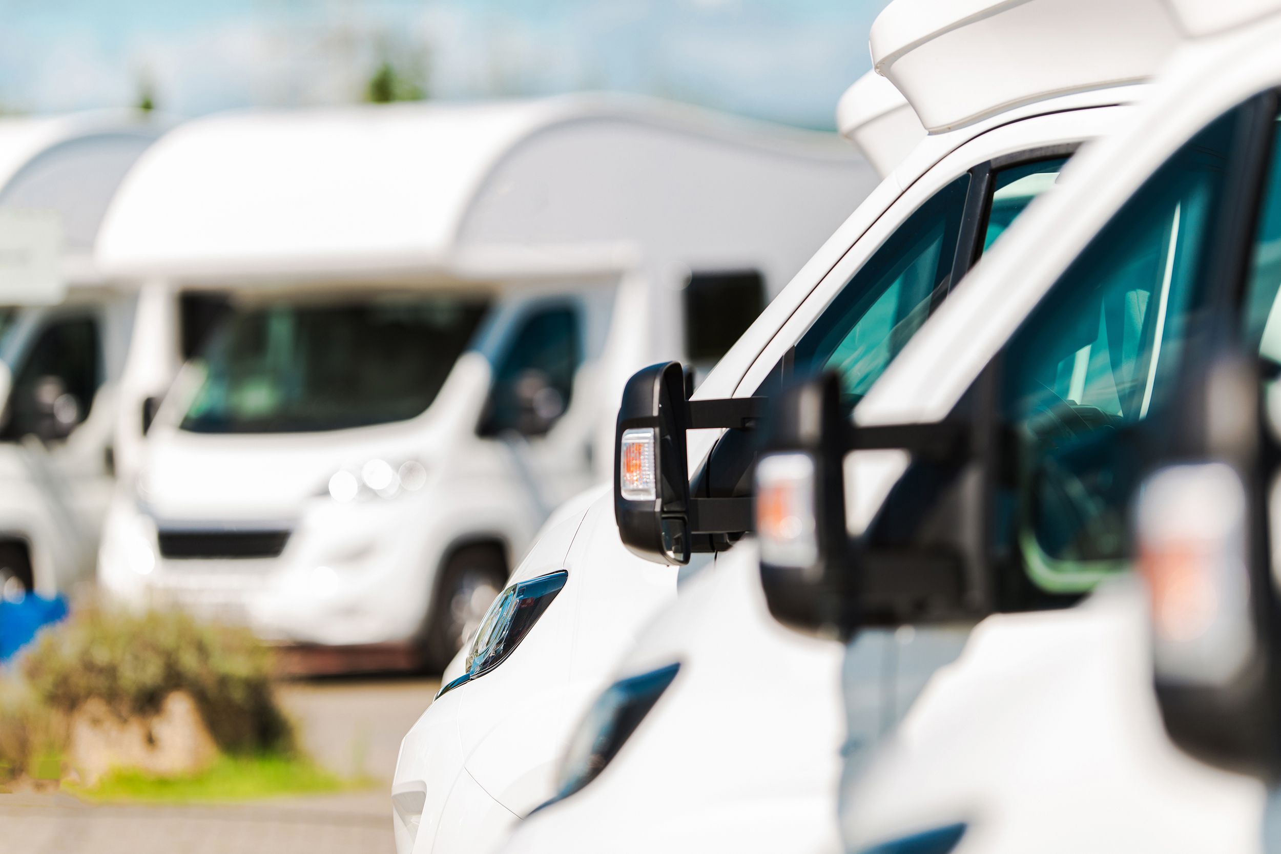 Closeup of front sides of a row of white RVs, selective focus of driver's windows, on the right foreground, on a dealership lot, two white RVs for sale, blurred, on the left background
