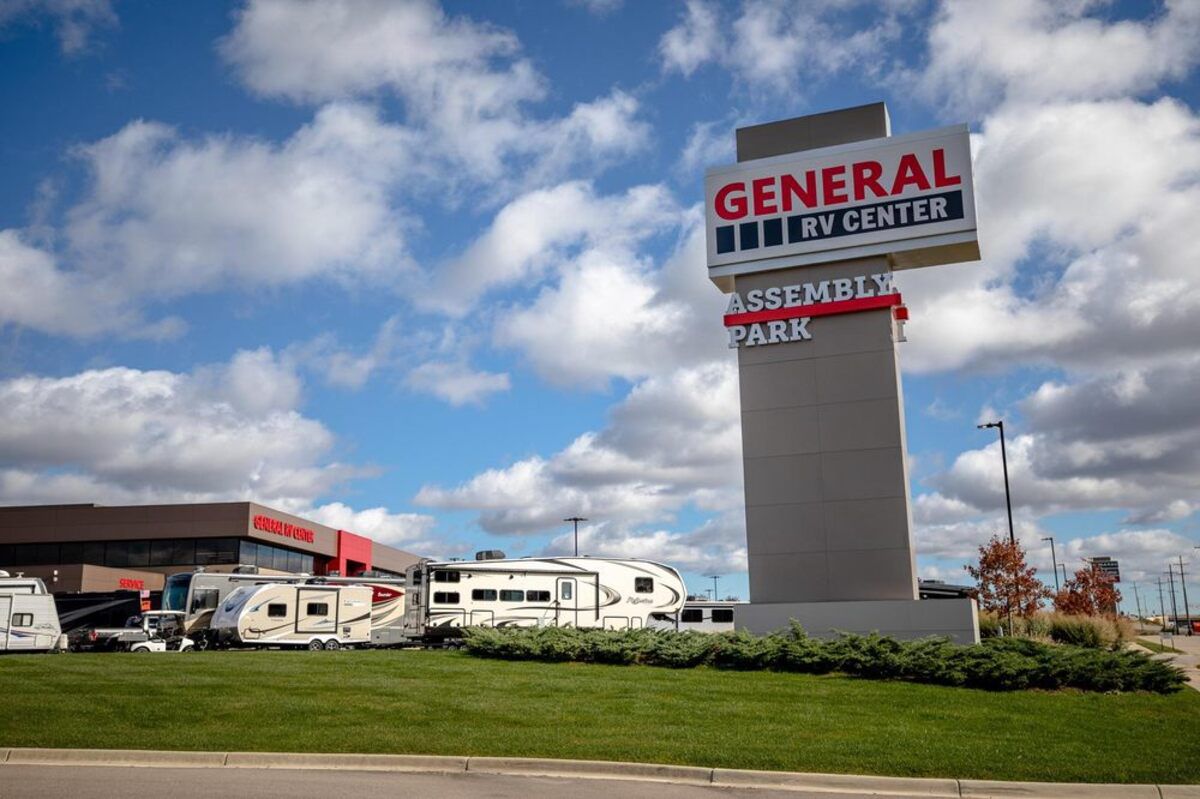 Exterior of General RV Center, Wixom, Michigan, as seen from the road, RVs for sale with a large sign in the foreground, building behind it, an expansive blue sky with white clouds in the background