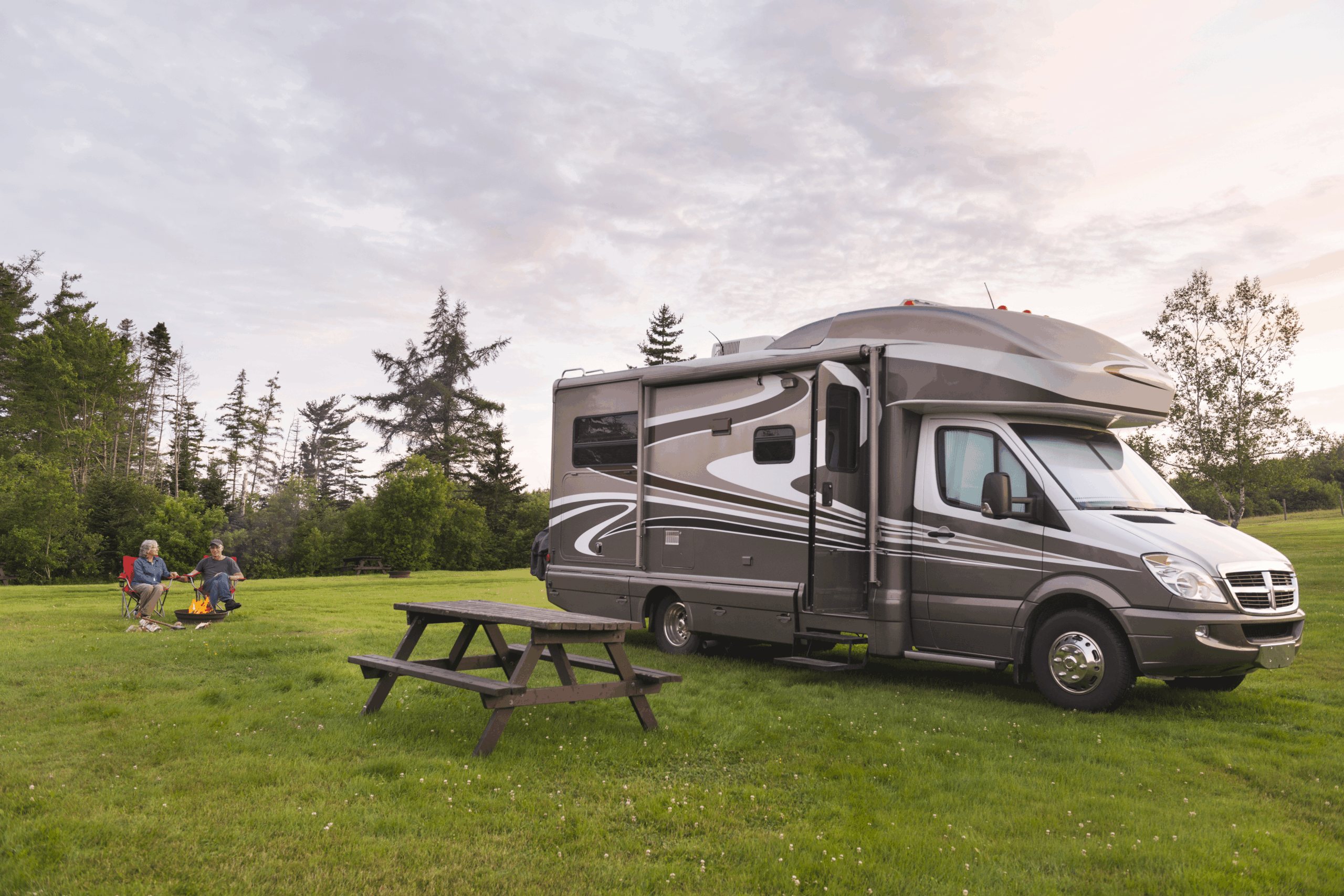 RV parked on grass near a wooden picnic table at a park in the foreground, couple seated in folding chairs with a campfire, forest and expansive sky during early sunset in the background