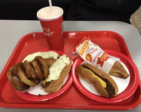 A red tray holds two hot dogs with slaw, a stack of onion rings, a chili cheese hot dog, a peach pie, and a large drink with a straw. All food is served on red and white plates on a grey tabletop.
