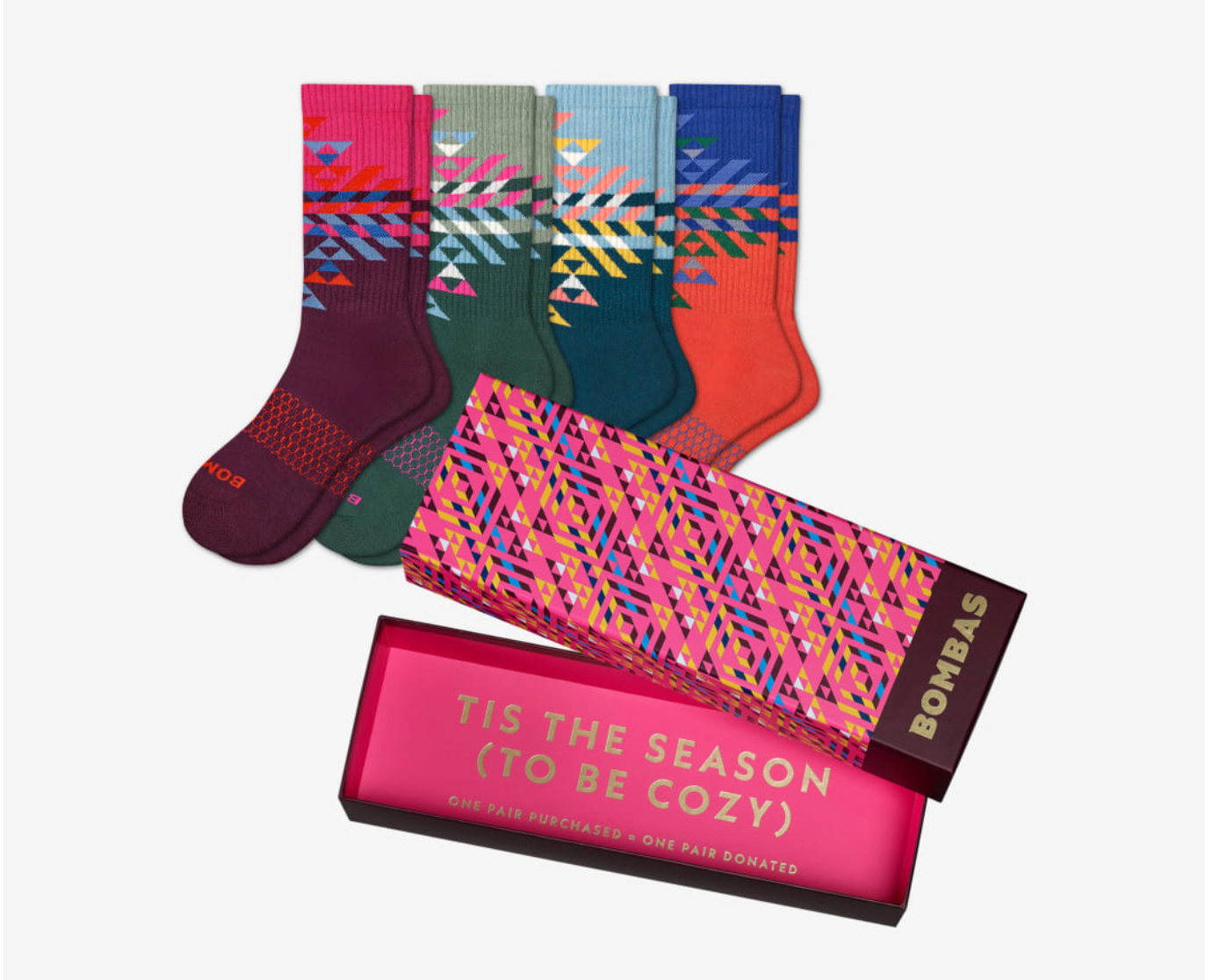 Four pairs of colorful, patterned socks are displayed next to an open festive Bombas gift box, which reads, “TIS THE SEASON (TO BE COZY)” inside the lid.