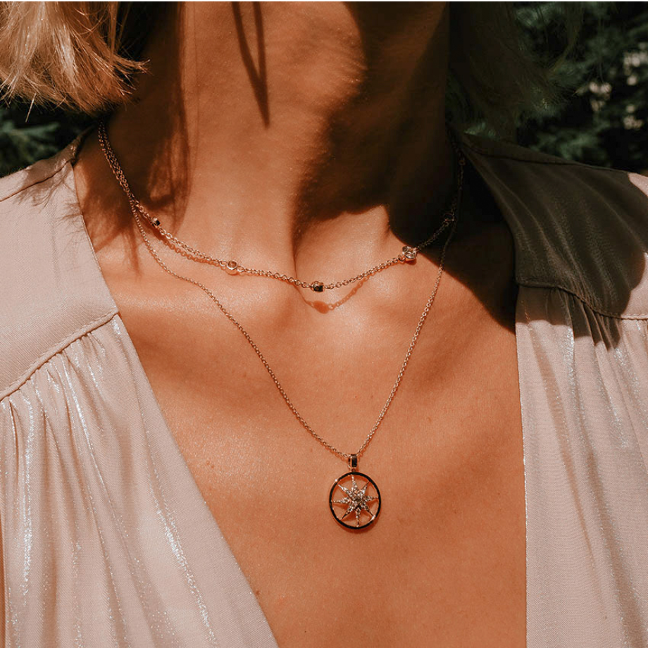 A woman wearing a v-neck beige blouse and two layered gold necklaces; the shorter necklace has small beads, while the longer one features a round pendant with a star design.