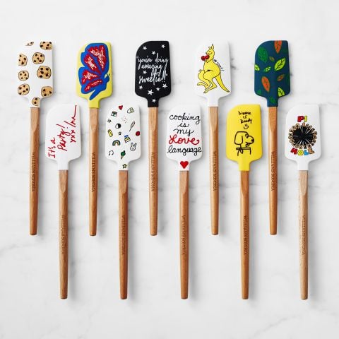 Nine colorful spatulas with wooden handles arranged on a white surface, each featuring a unique decorated design such as cookies, butterflies, flowers, animals, and playful hand-lettered messages.