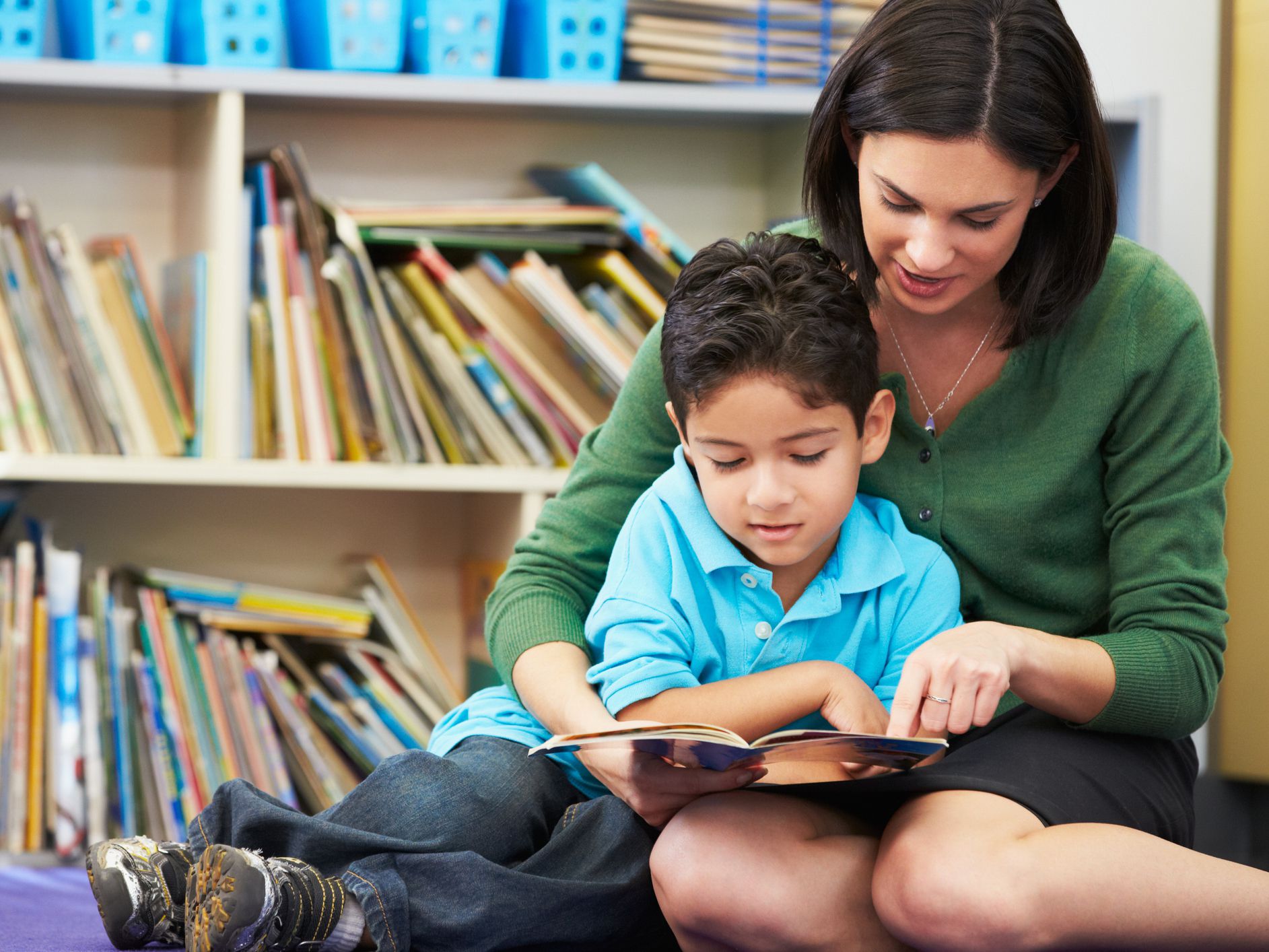 Elementary Pupil Reading With Teacher In Classroom Sitting Down On Floor