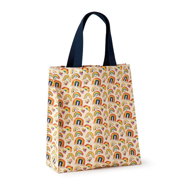 A reusable tote bag with navy blue handles and a colorful pattern featuring rainbows, sunshine, hearts, and flowers on a light background.