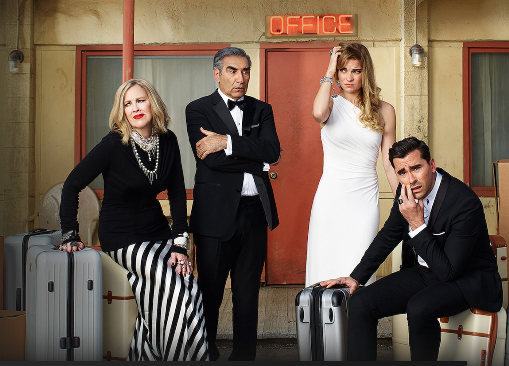 Four people dressed formally sit and stand with suitcases outside a motel office. Two are seated, one woman in a black and white outfit and one man in a tuxedo. Another woman in white and a man in black sit and stand beside them.