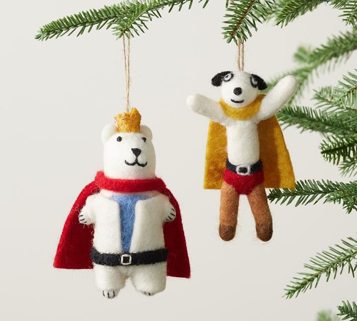 Two felt animal ornaments, a bear with a crown and cape and a dog with a yellow cape and red shorts, hang from a green pine branch. The background is plain and light-colored.