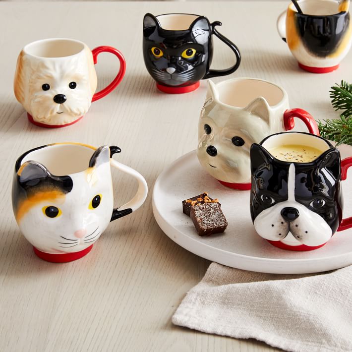 Five animal-themed mugs shaped like cats and dogs are arranged on a light table with a plate holding a drink and small desserts. A beige napkin and a small green plant are also visible.