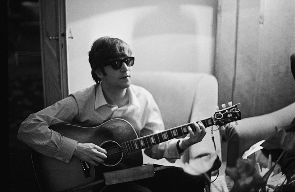 John Lennon (1940 - 1980) of the Beatles plays the guitar in a hotel room in Paris,